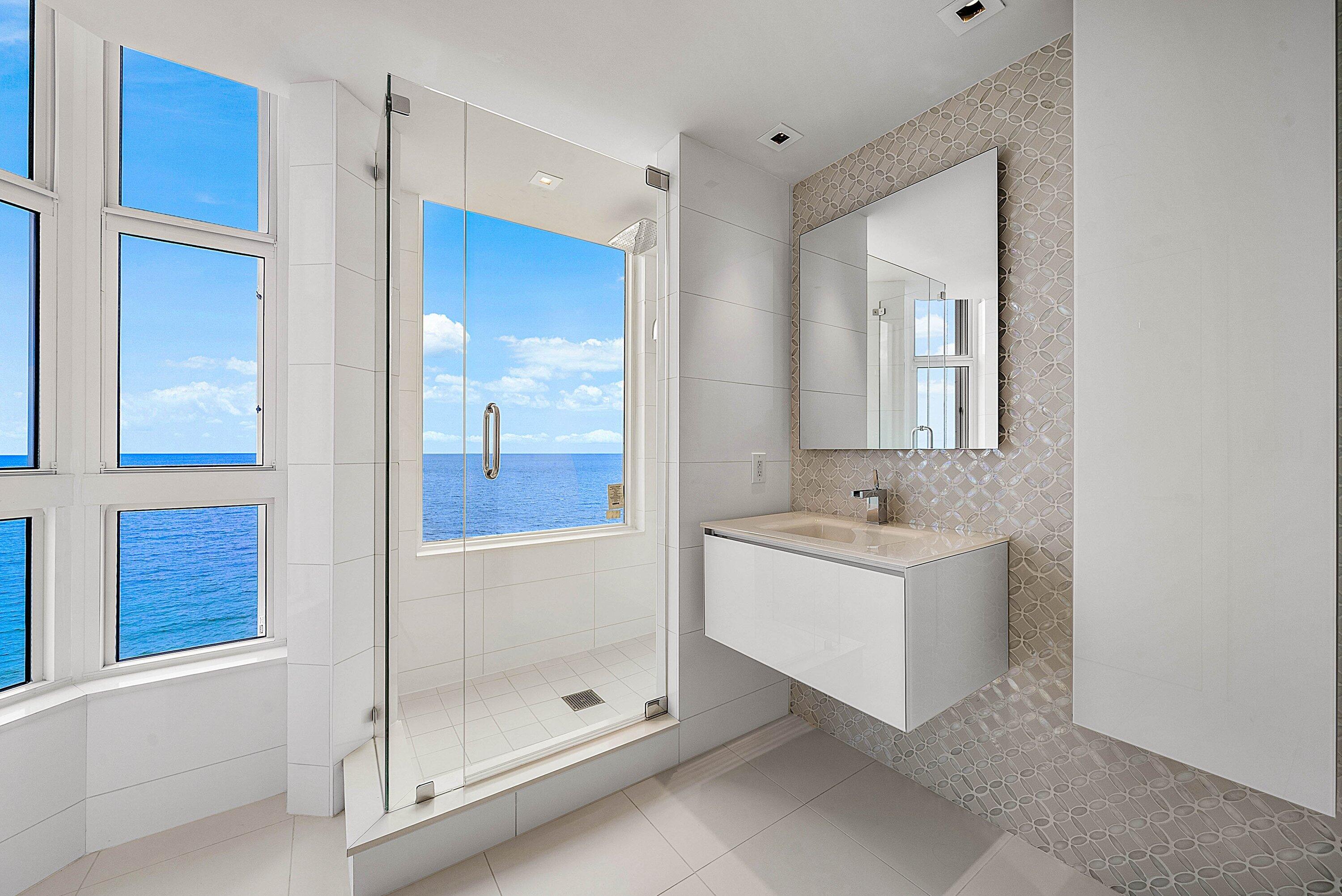 Glass-enclosed shower with integrated window, floating vanity, wall-mounted mirror, mosaic tile accent wall, and floor-to-ceiling windows with water backdrop