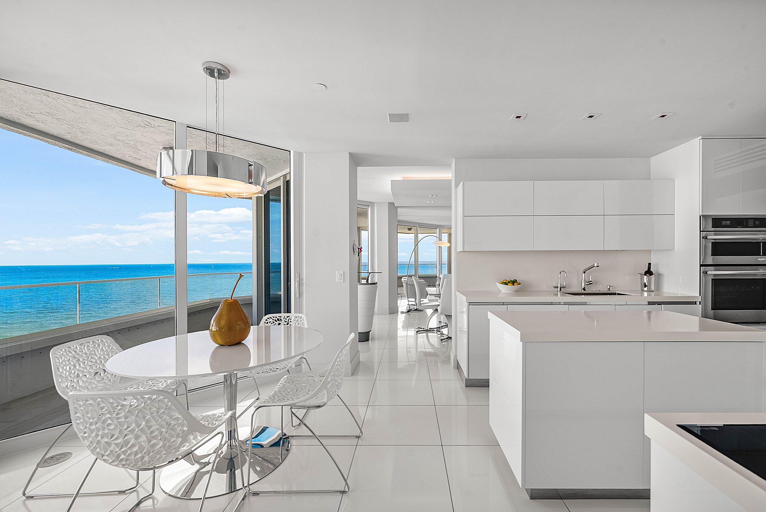 Floor-to-ceiling glass paneling, high-gloss white cabinetry, center island with stone countertop, large-format tile flooring, and integrated stainless steel double wall ovens