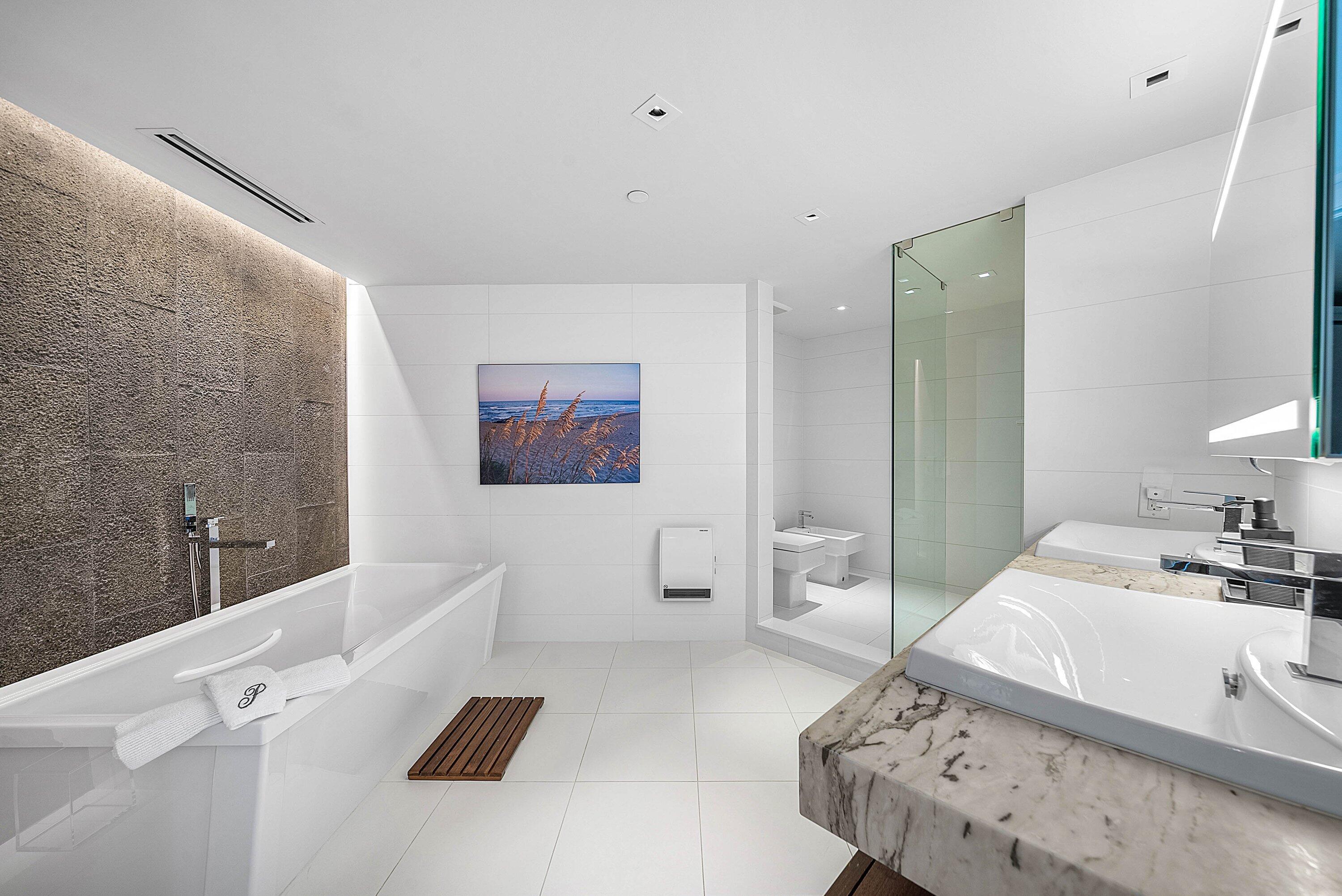 Bathroom featuring a freestanding white soaking tub, textured stone accent wall, dual-sink vanity with stone countertop, frameless glass-enclosed shower, and large-format tile flooring
