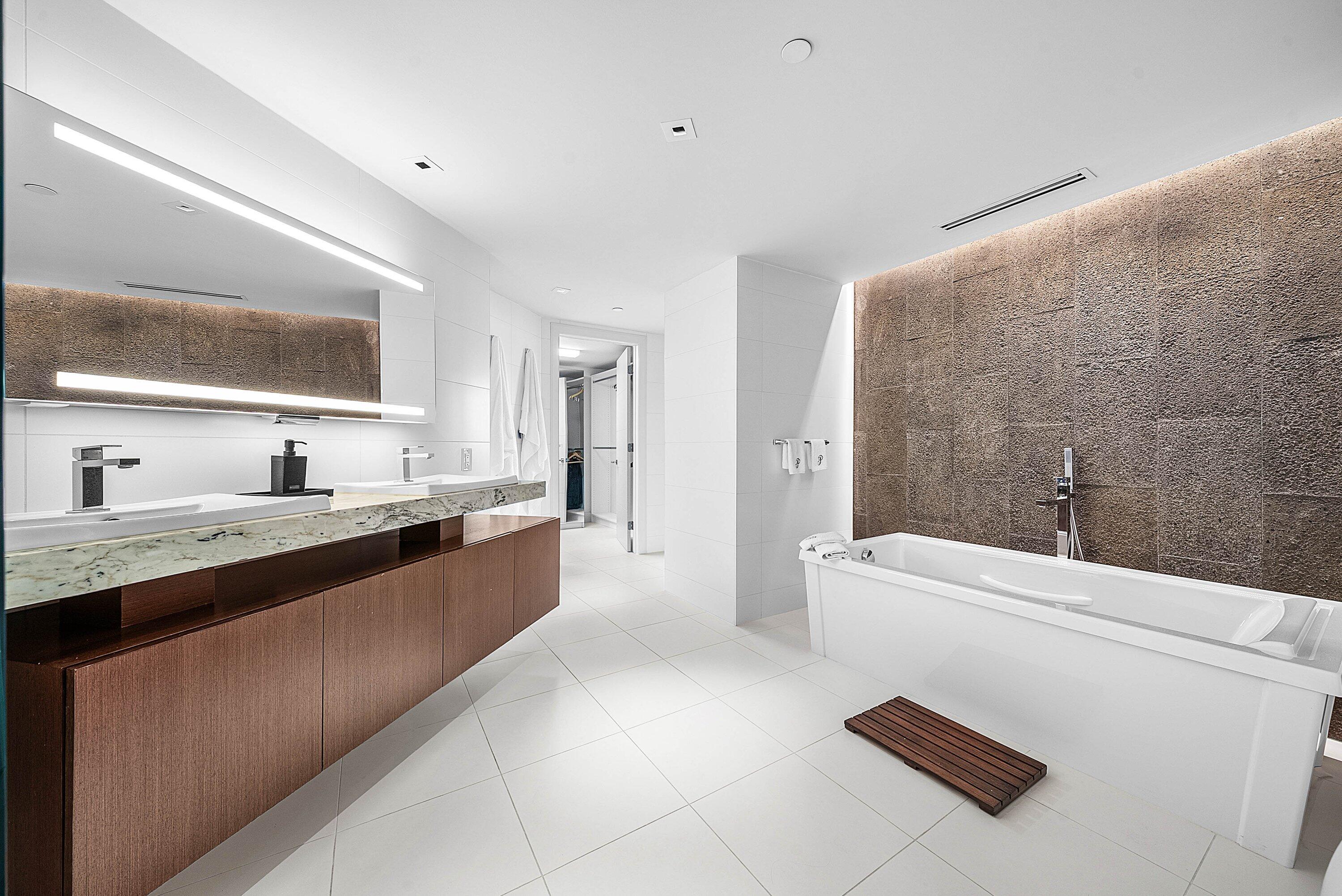 Floating wood-finish double vanity with stone countertop, large LED-backlit mirror, soaking tub, textured stone feature wall, and recessed ceiling lighting