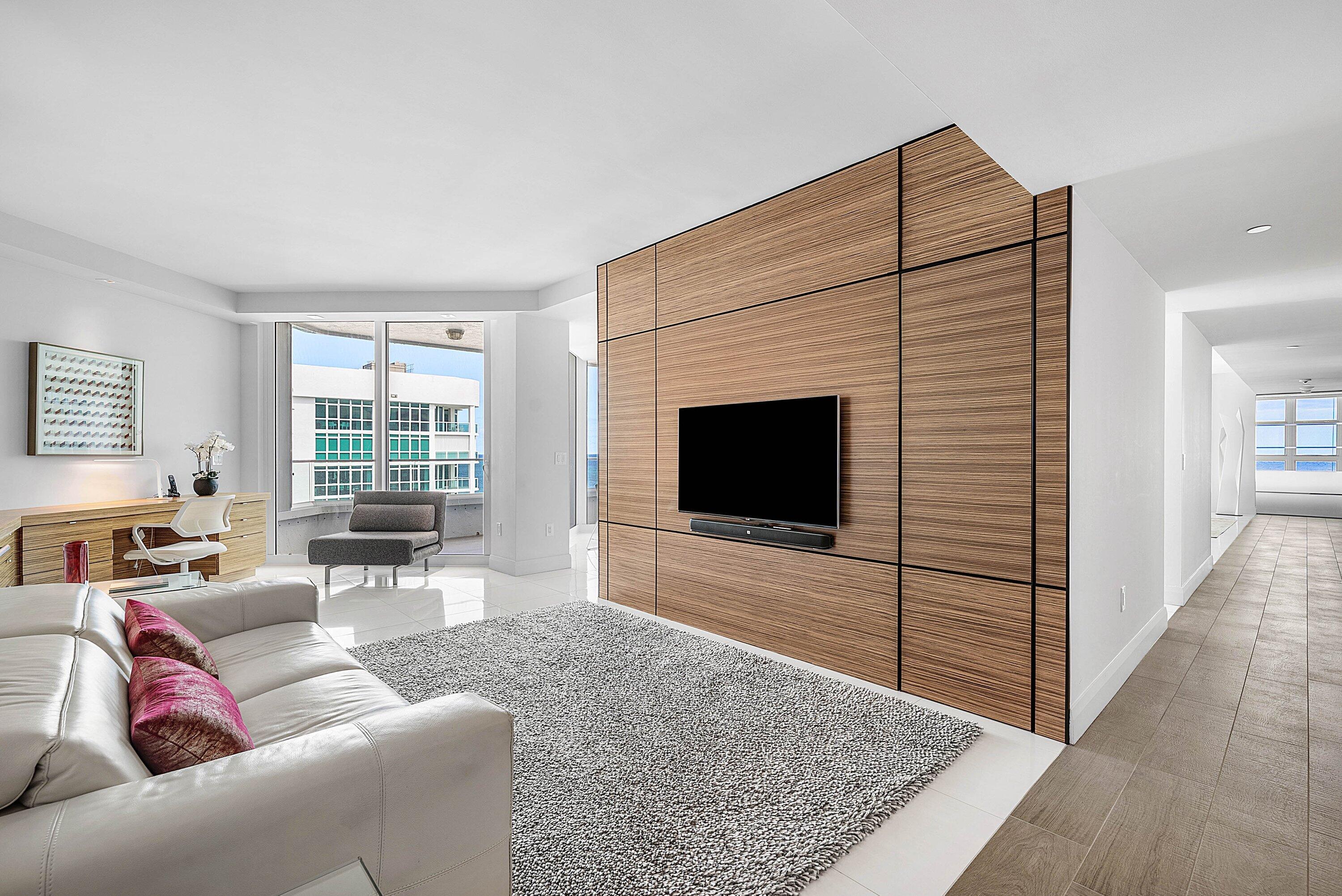 Custom floor-to-ceiling wood-panel accent wall with integrated television mount, expansive floor-to-ceiling glass doors, and polished stone flooring