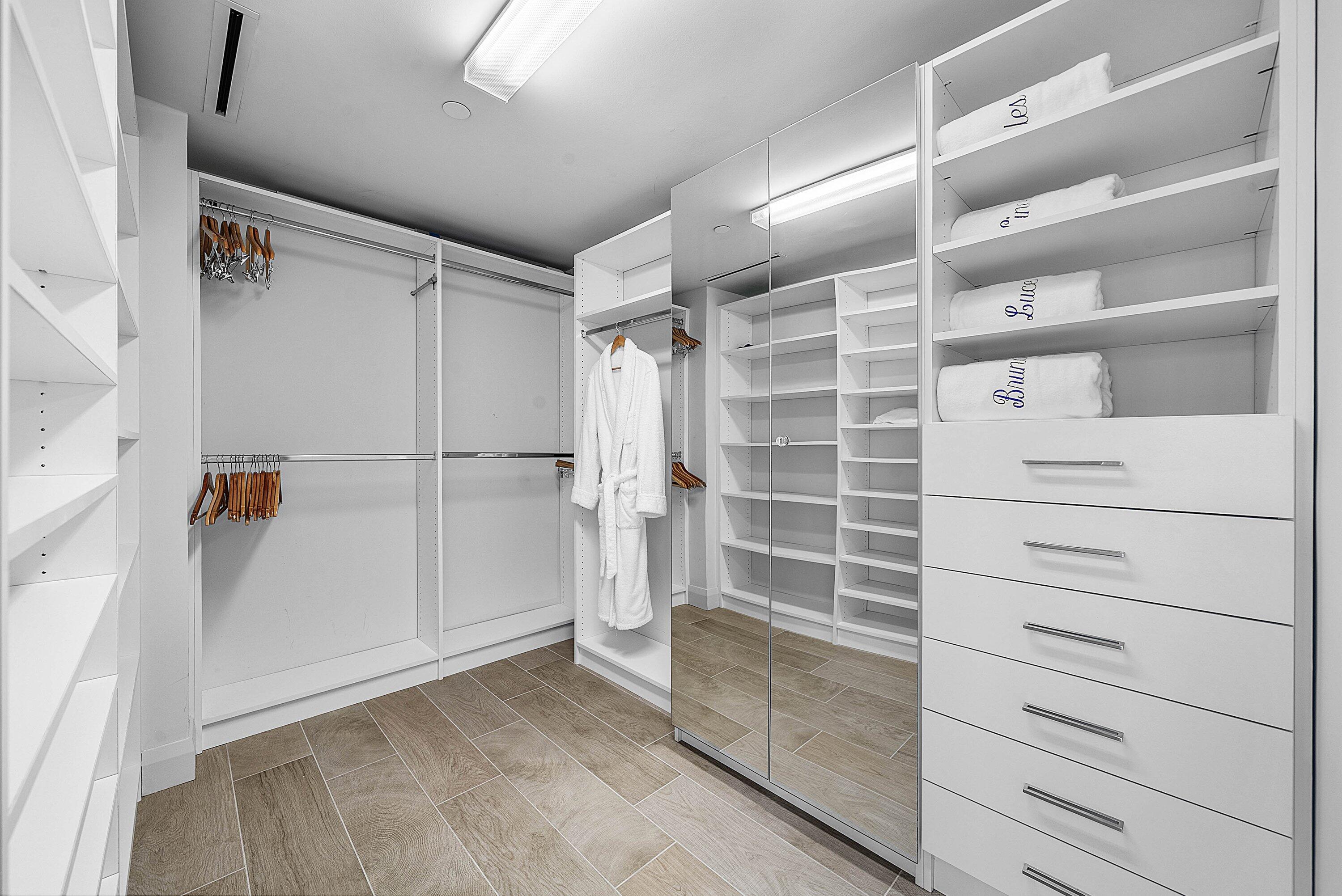 Custom storage system featuring floor-to-ceiling white cabinetry, multi-level hanging rods, built-in shelving, integrated mirror panels, and wood-finish tile flooring