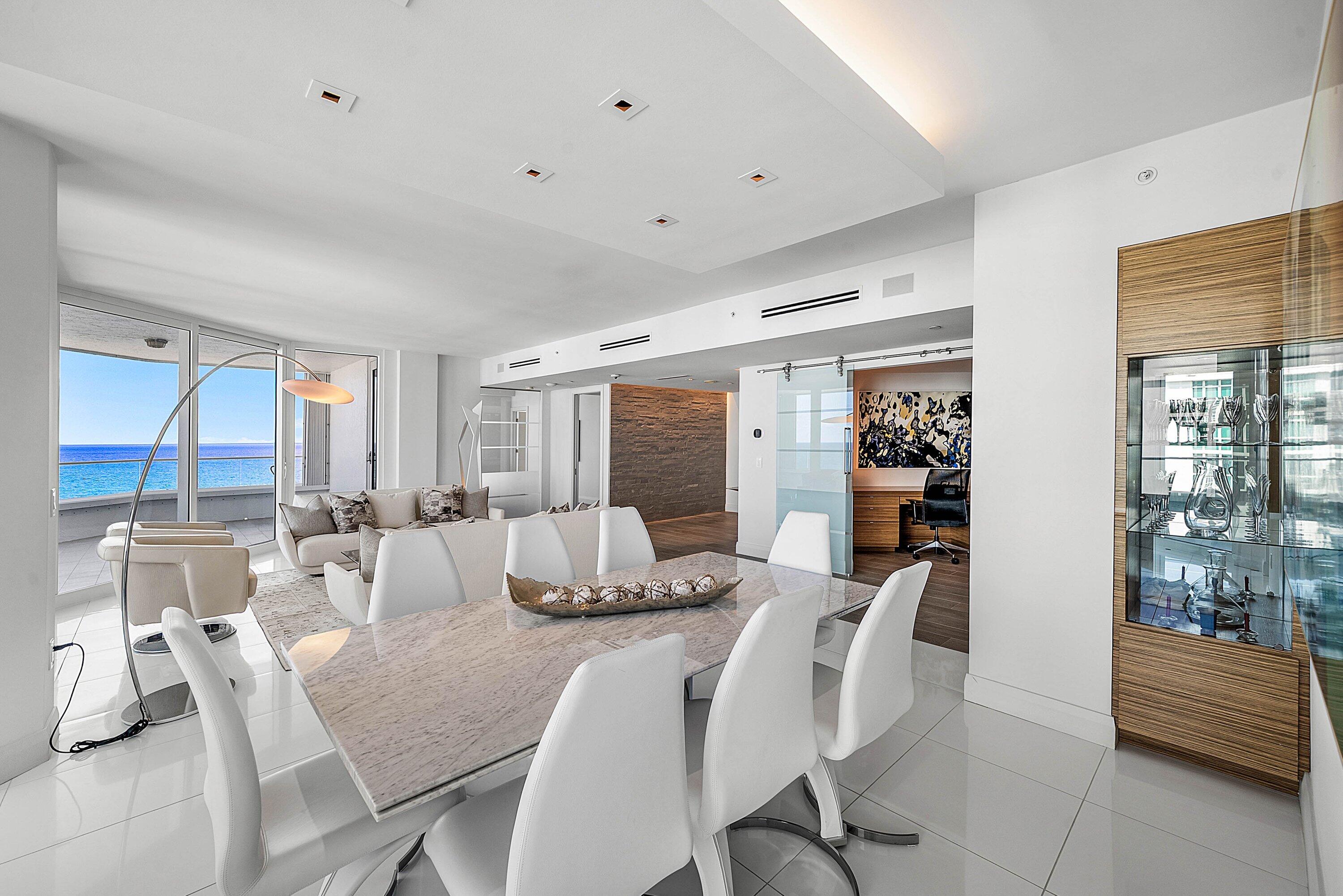 Open-concept living space featuring high-gloss tile flooring, recessed tray ceiling lighting, built-in wooden cabinetry with glass-front shelving, sliding glass door room entry, and floor-to-ceiling windows with coastal horizon scenery