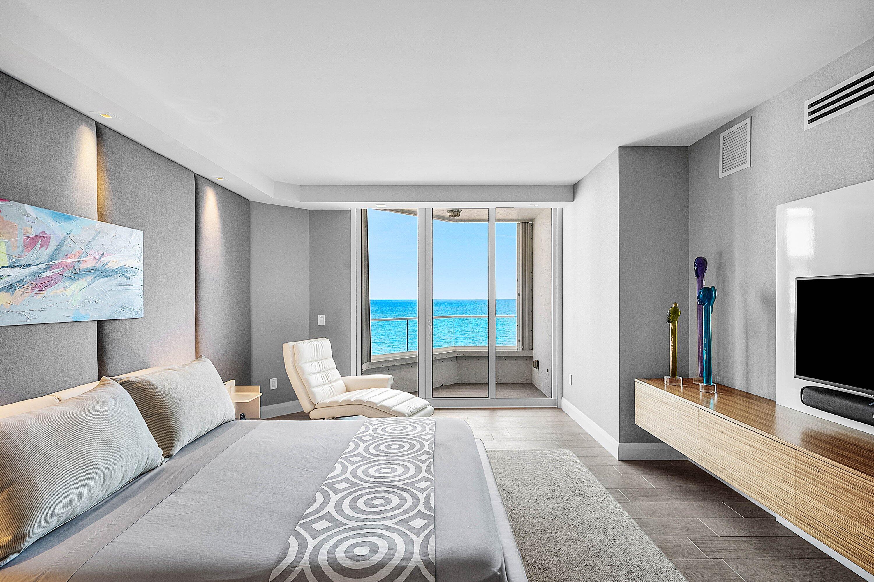 Floor-to-ceiling glass sliding doors opening to a balcony, wall-to-wall upholstered headboard paneling, wall-mounted wood-finish media console, and large-format tile flooring