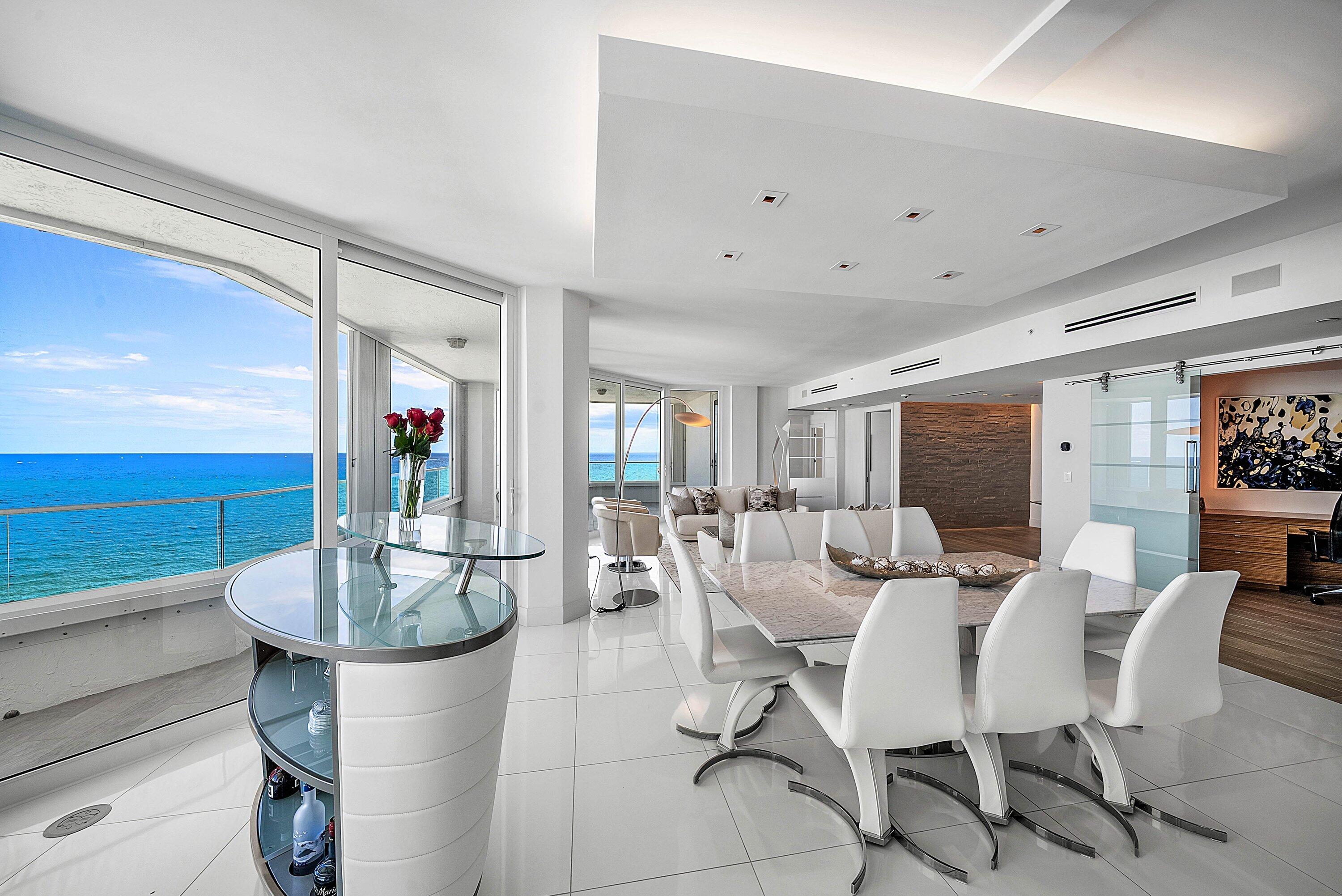 Expansive floor-to-ceiling windows, polished tile flooring, tiered tray ceiling with recessed lighting, integrated glass and stone-finish bar, and sliding frosted glass partition