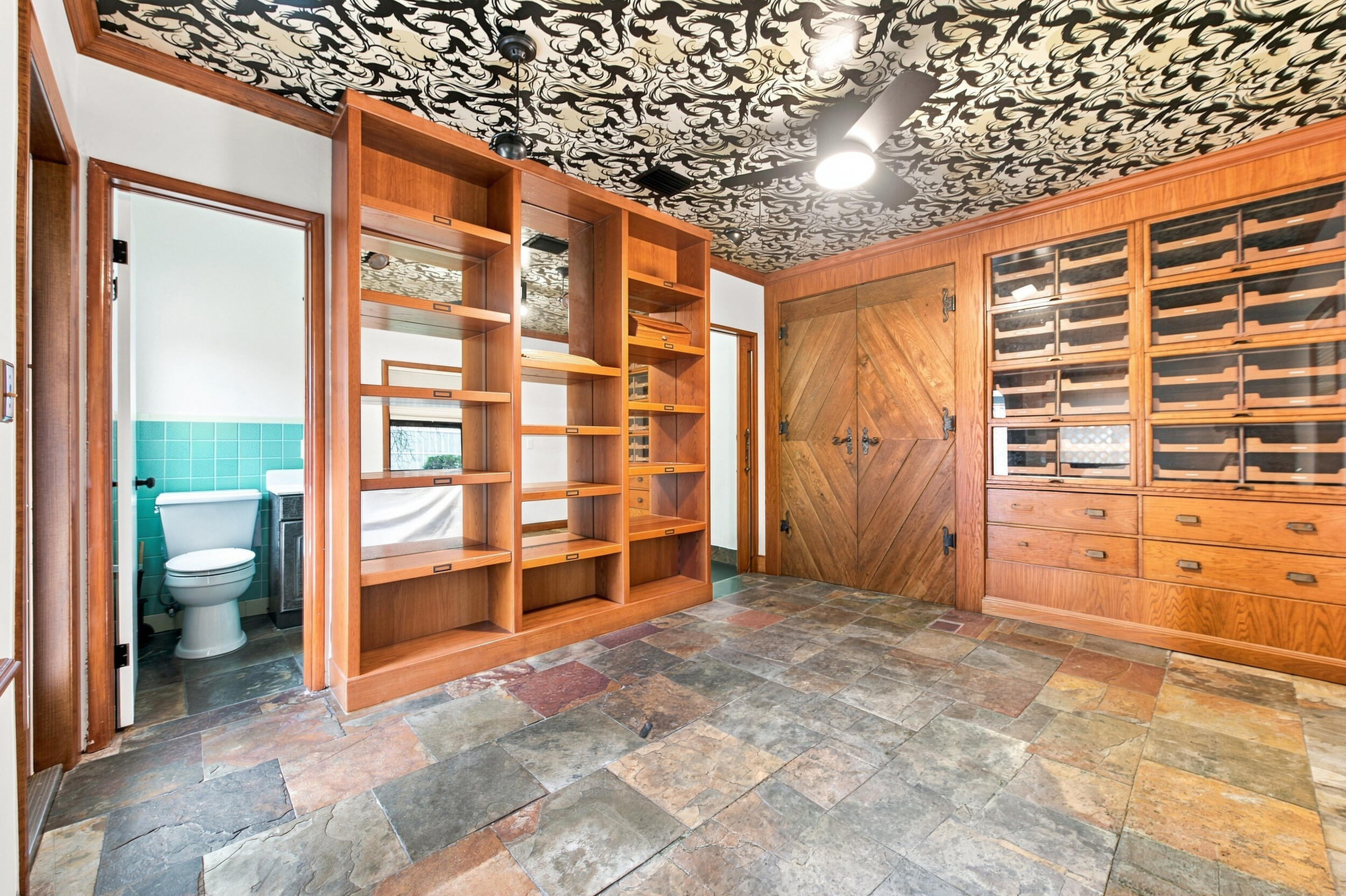 Custom wood cabinetry with integrated shelving and glass-front storage, patterned ceiling, multi-tonal stone tile flooring, and double doors featuring a diagonal wood plank design
