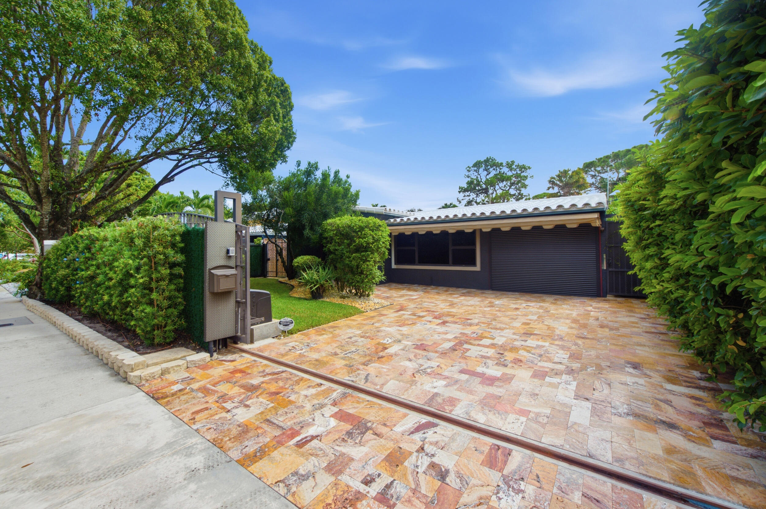 Herringbone-patterned stone driveway, gated entry, mature privacy hedging, tile roof, and enclosed garage