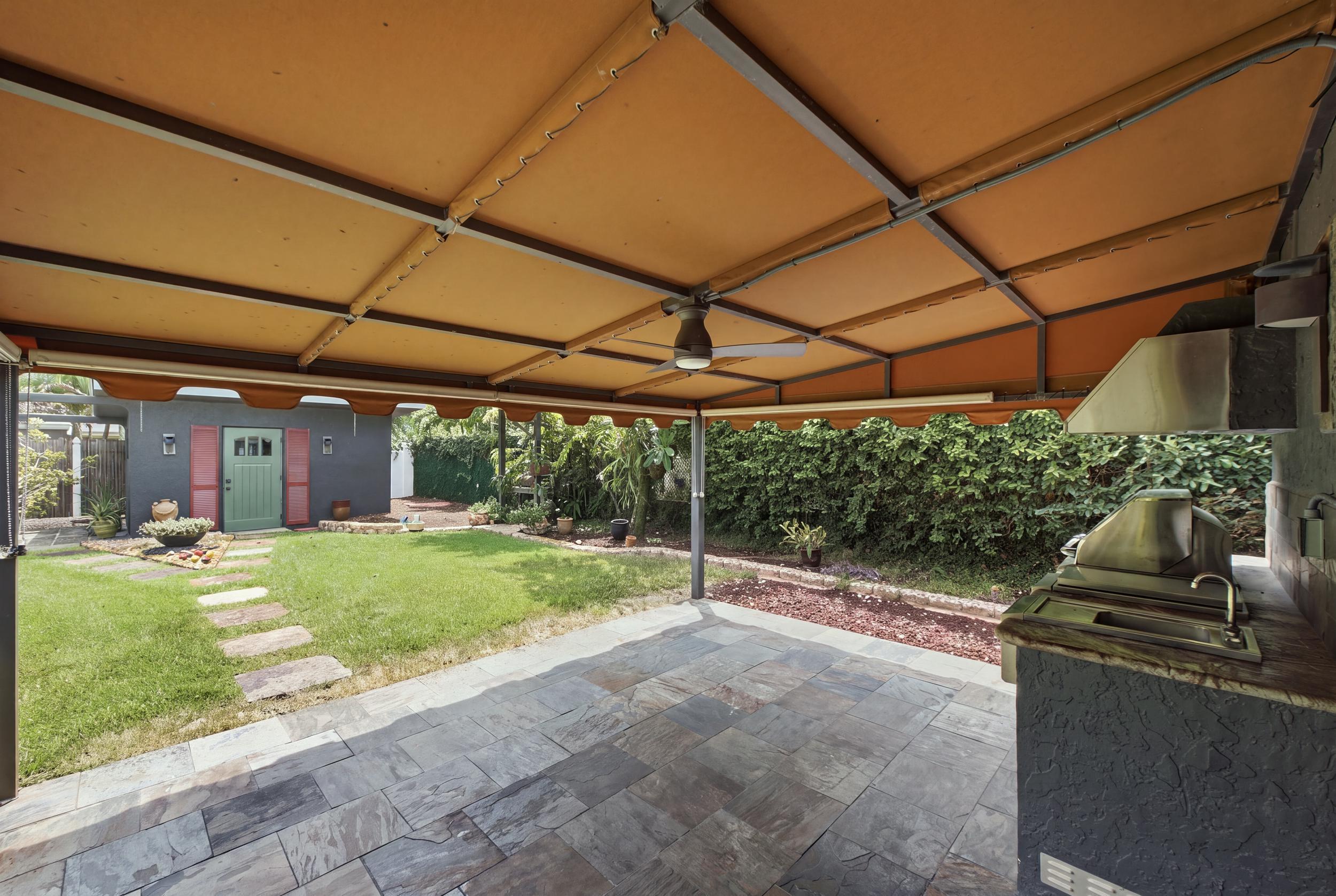 Covered outdoor patio featuring stone tile flooring, a built-in outdoor kitchen with stainless steel grill and sink, and a ceiling fan