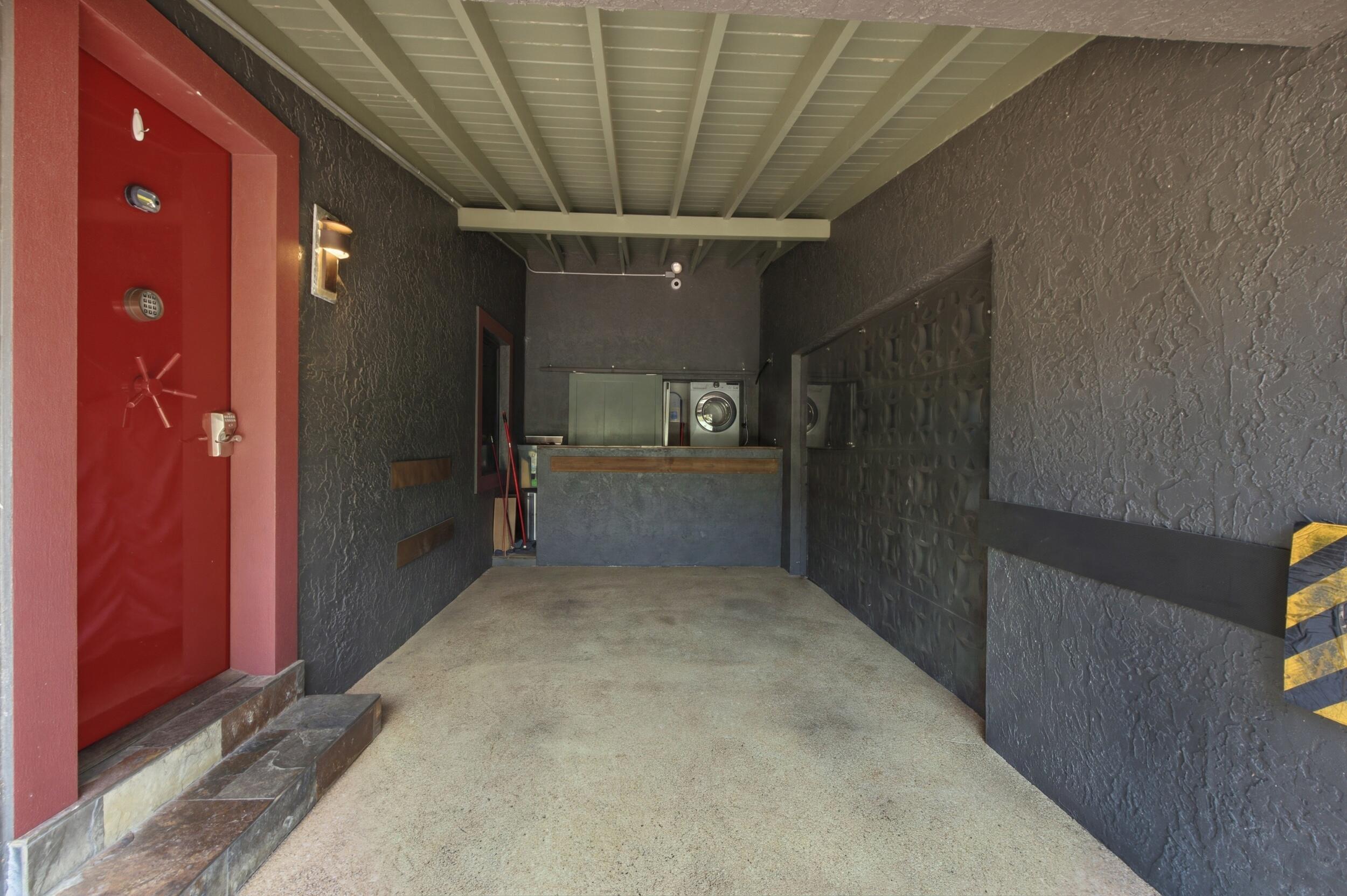 Covered carport featuring a vibrant red entry door, textured dark wall cladding, concrete flooring, and an elevated storage platform