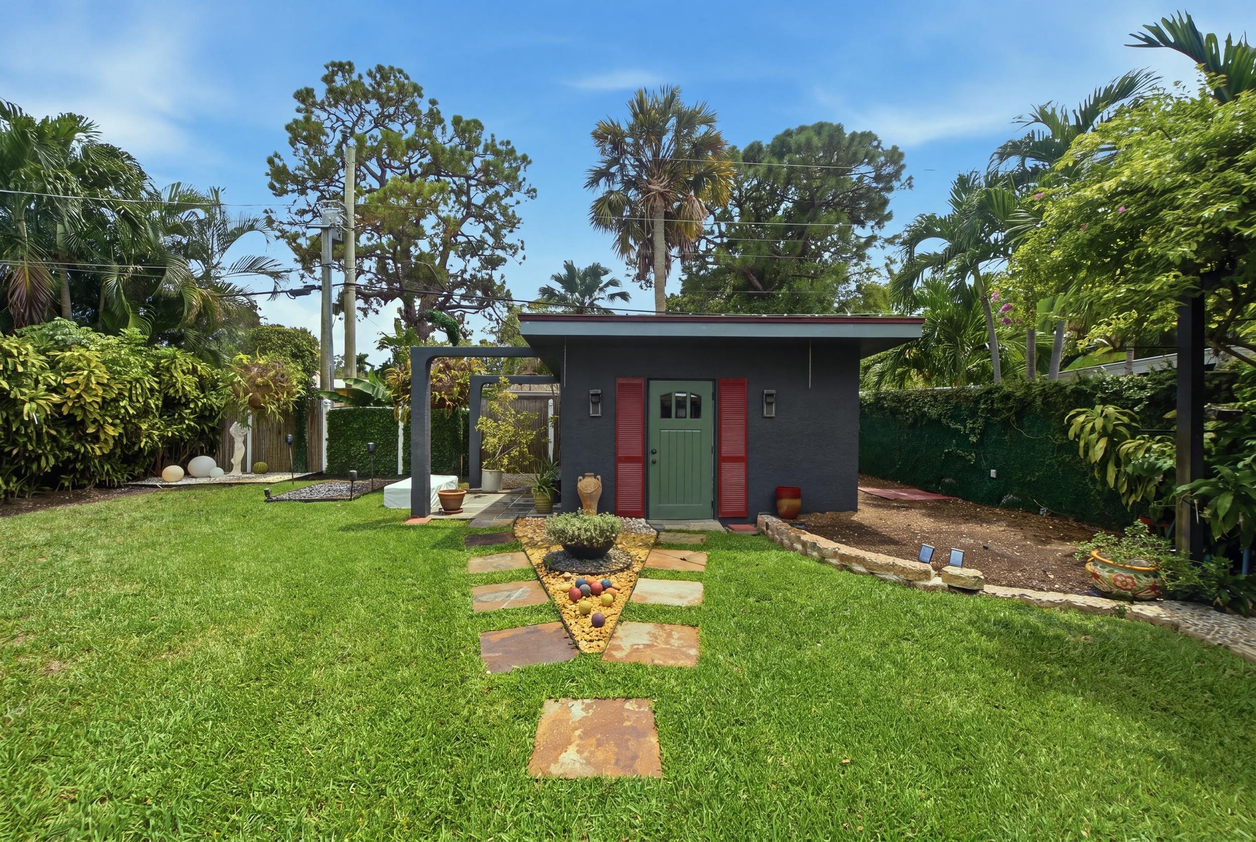 Detached structure featuring a masonry exterior, decorative red shutters, a green paneled entry door, a stone-paved walkway, and mature landscaping