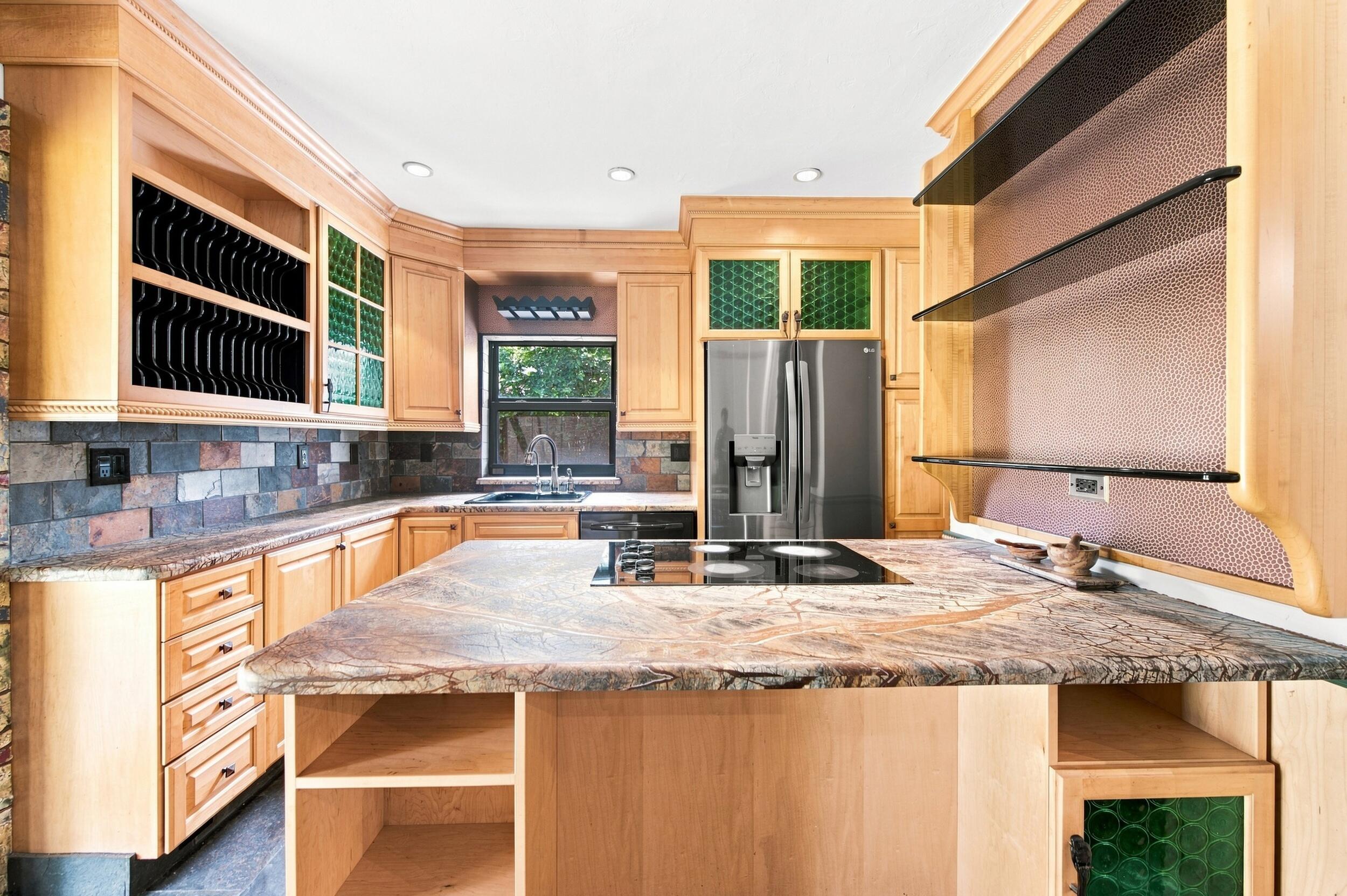 Chef's kitchen featuring custom wood cabinetry with glass-front upper cabinets, a large center island with stone countertop, stainless steel appliances including a French-door refrigerator, tile backsplash, and abundant recessed lighting throughout