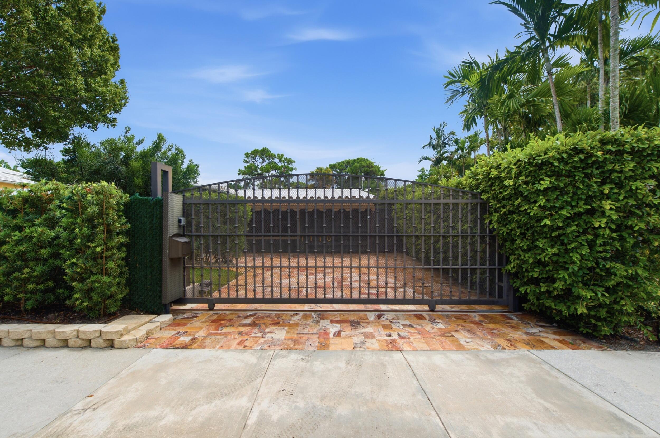 Private entrance featuring a motorized wrought iron gate, stone-paved driveway, and perimeter hedges