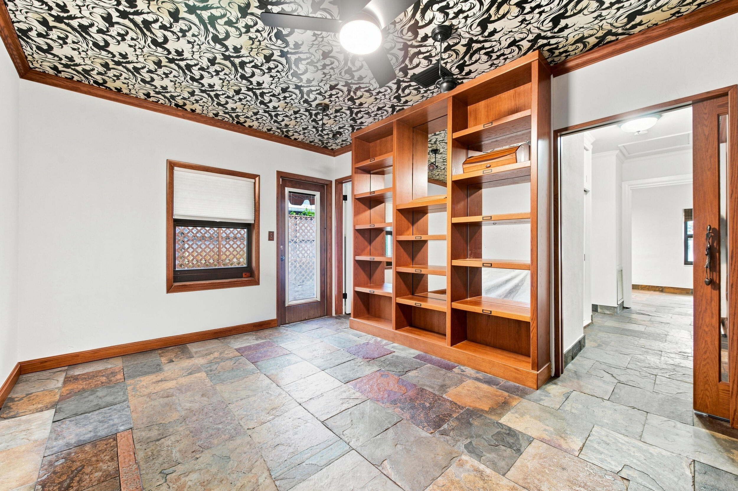 Room featuring natural stone tile flooring, floor-to-ceiling built-in wood shelving, intricate patterned ceiling treatment, and wood-trimmed window and door frames