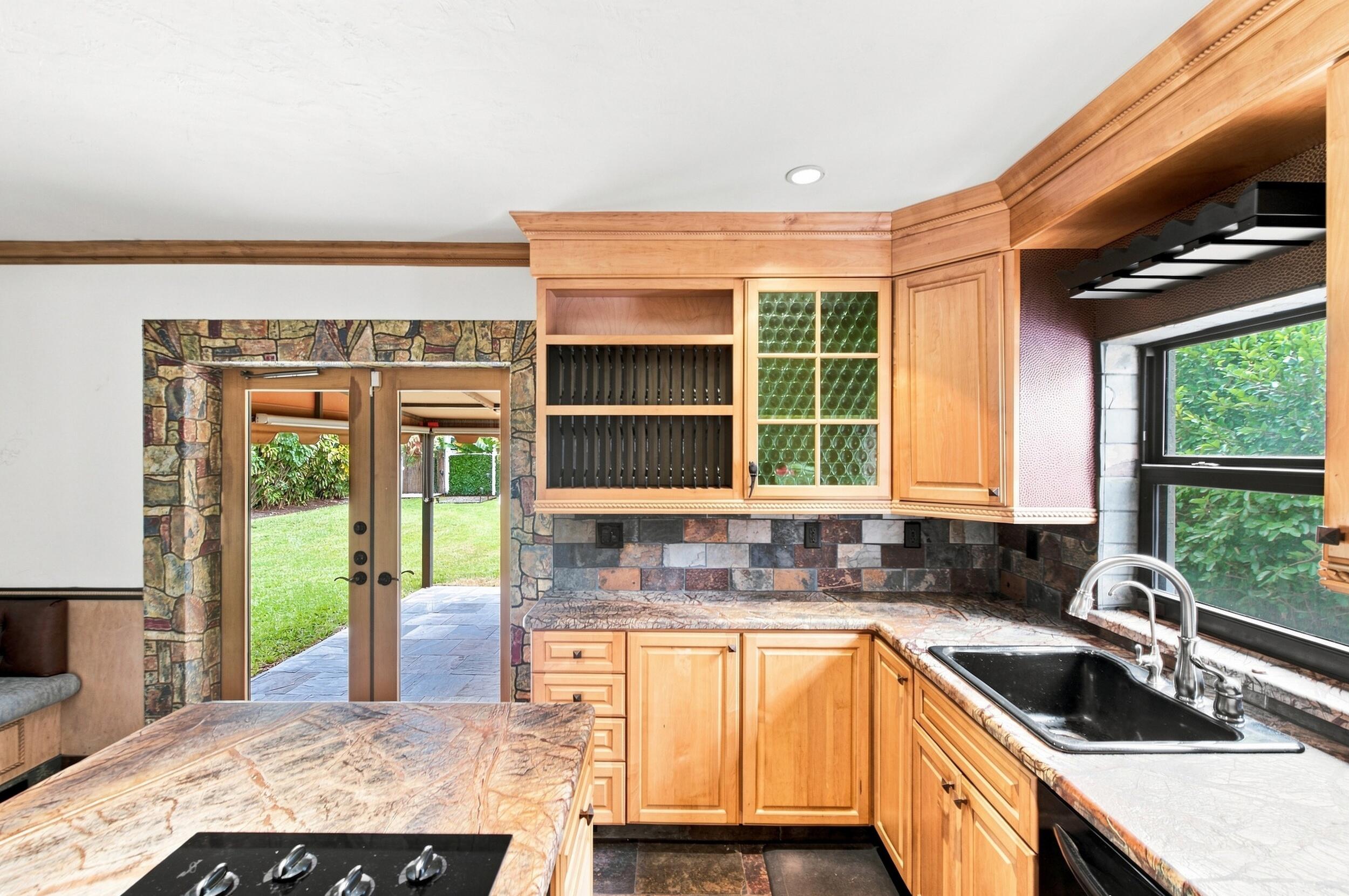 Kitchen featuring custom natural wood cabinetry, stone tile backsplash, integrated stone-topped island with stovetop, drop-in sink, and double glass doors encased in stone masonry