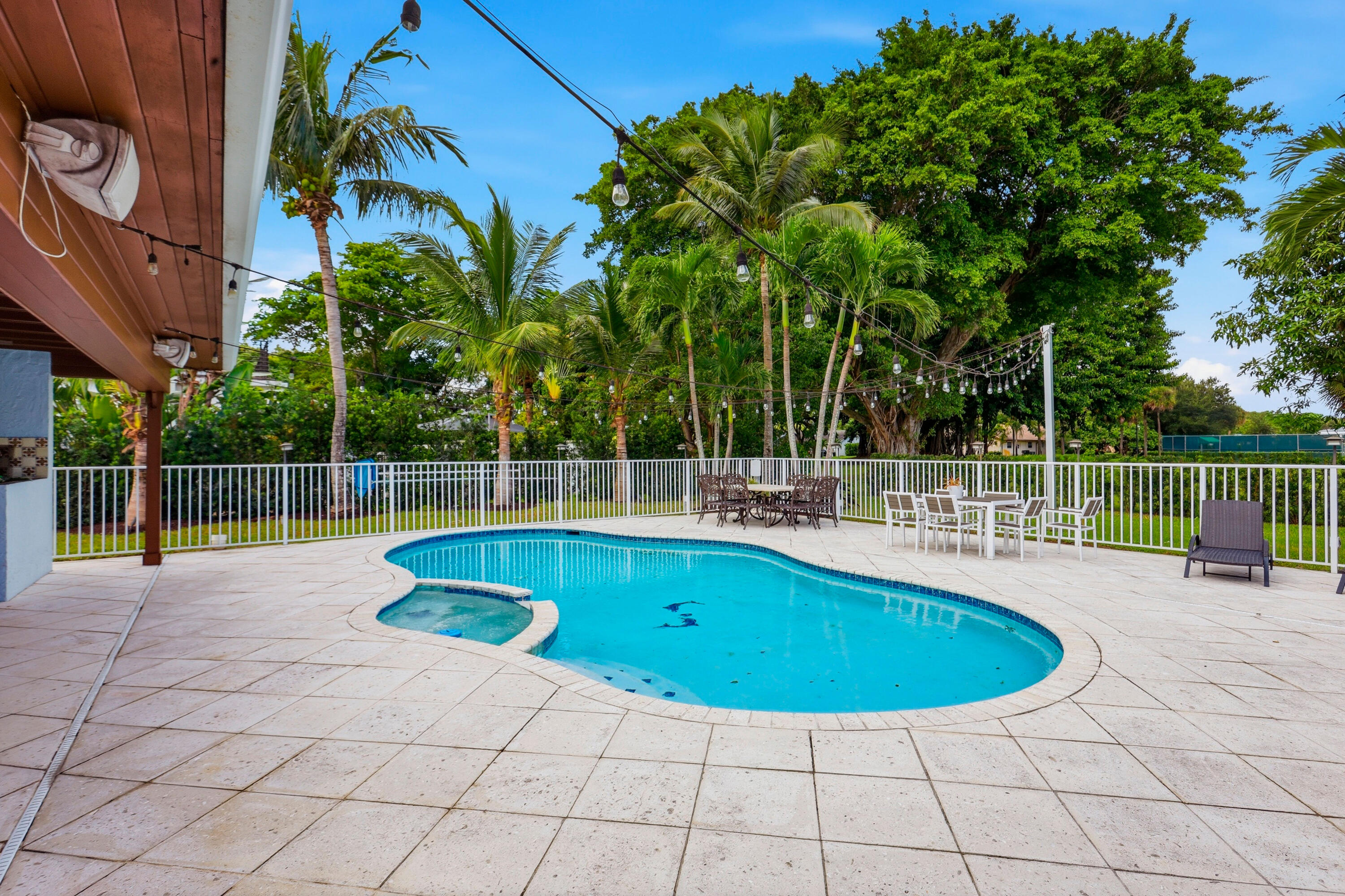 Outdoor living area featuring a custom-shaped swimming pool with integrated spa, expansive stone paver patio, covered porch with wood-finish ceiling, and perimeter metal fencing