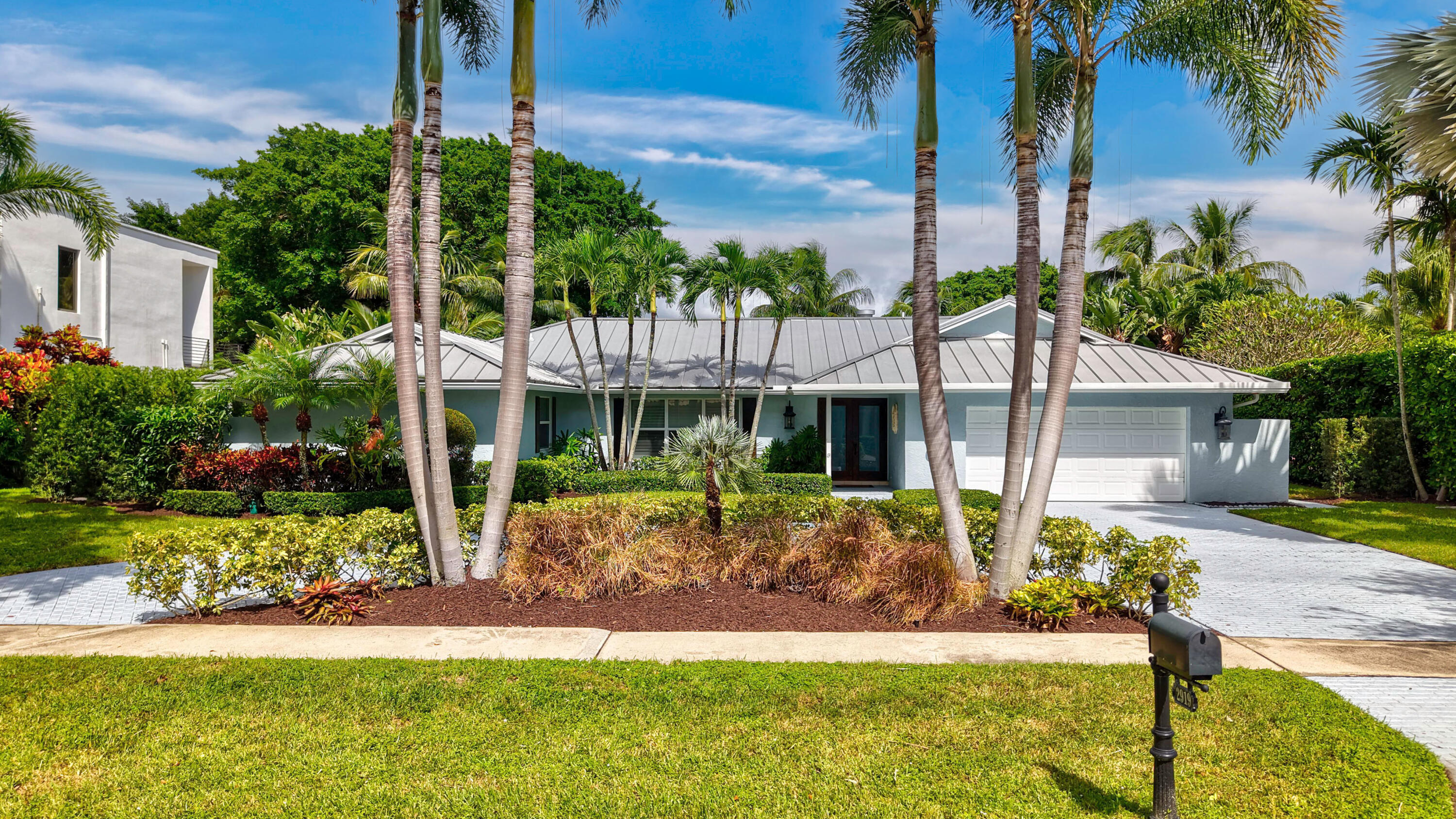 Single-story residence featuring a standing-seam metal roof, attached garage, stone-paved driveway, and mature palm trees