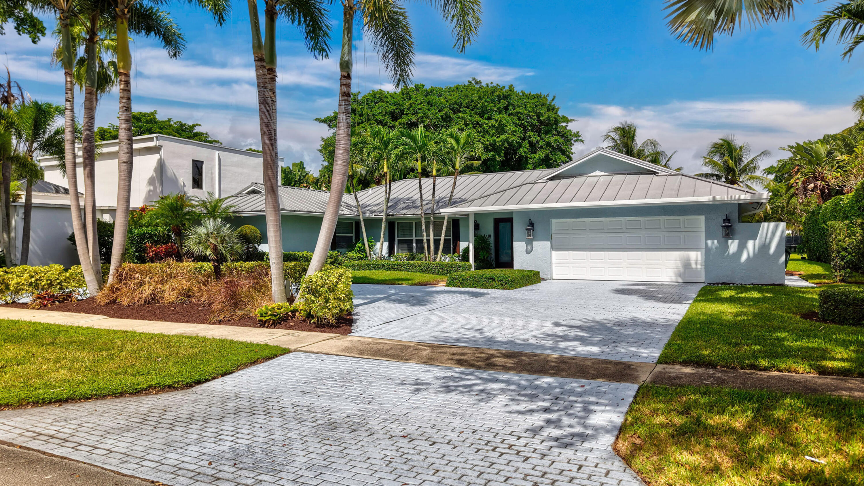 Single-story residence featuring a standing seam metal roof, paved driveway, two-car garage, and established tropical landscaping