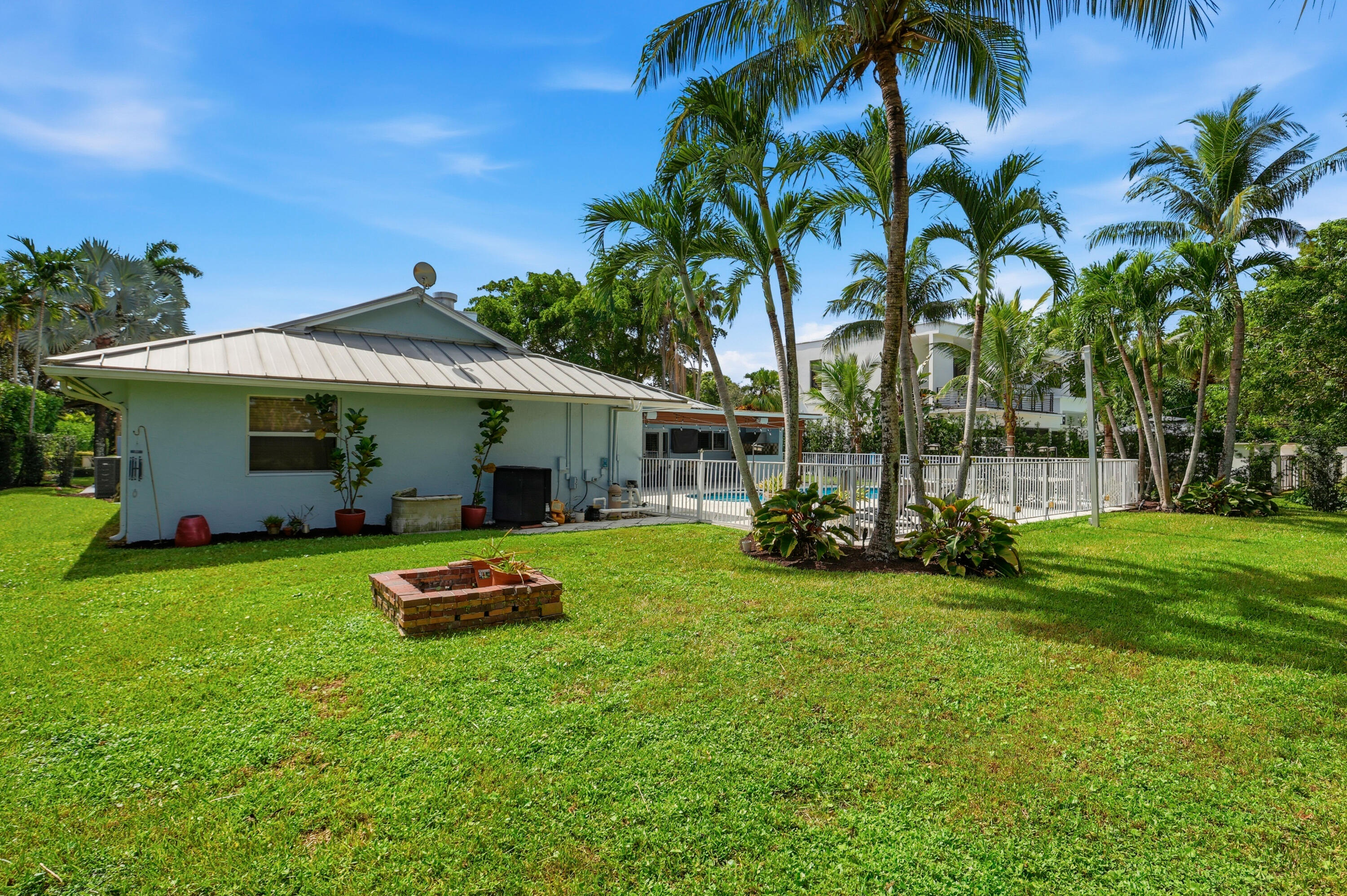Expansive backyard featuring a metal roof structure, a masonry fire pit, mature palm trees, and a fenced swimming pool area