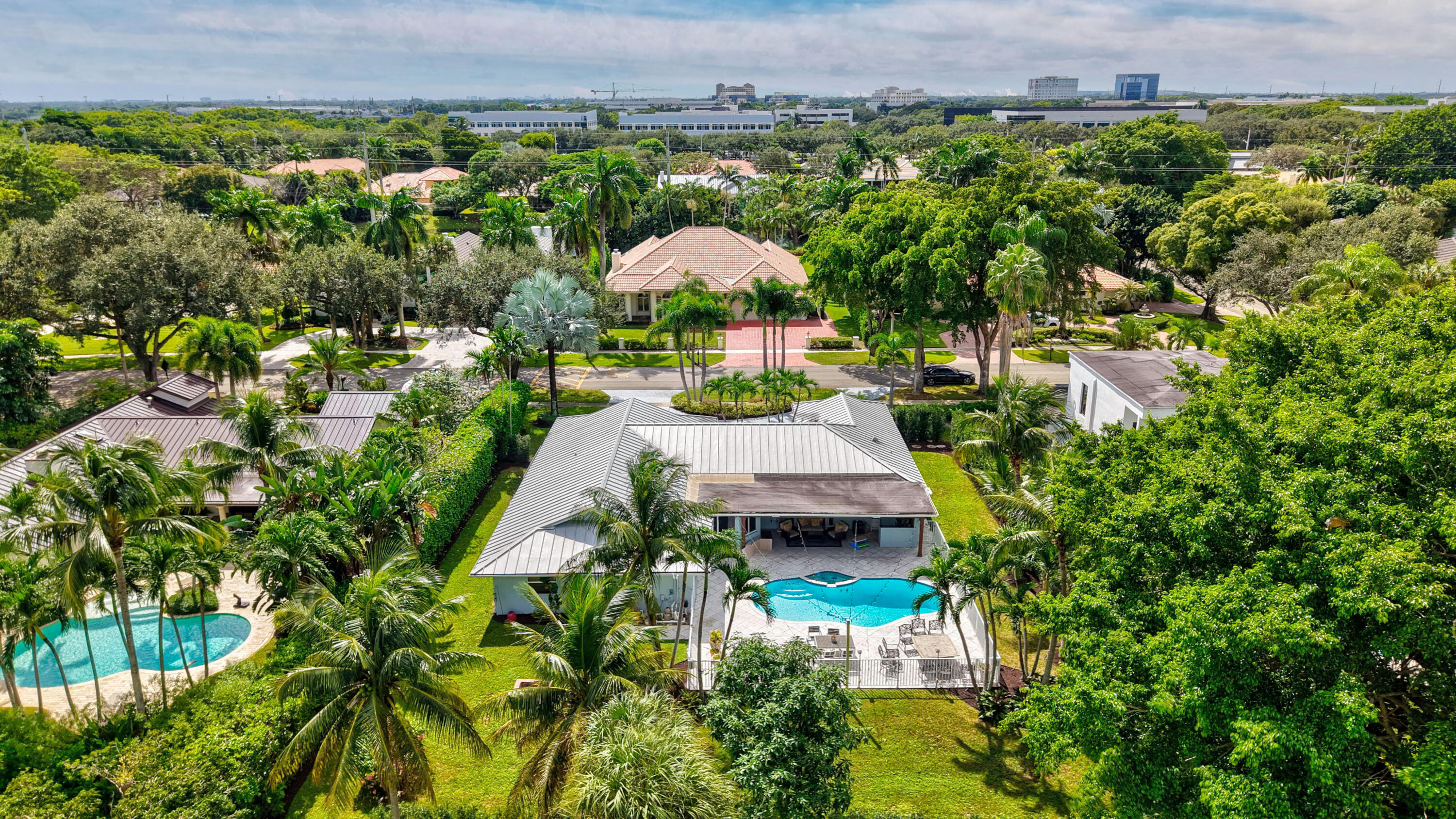 Aerial perspective featuring a single-story residence with a metal roof, an in-ground swimming pool, a stone-paved patio, and mature tropical landscaping