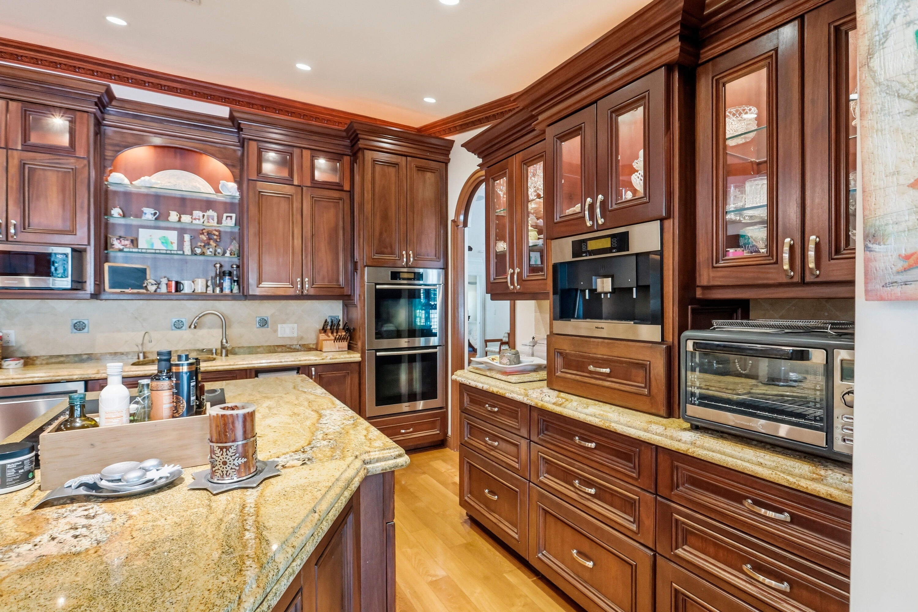 Custom dark-wood cabinetry with glass-panel upper doors, stone countertops, double wall ovens, integrated built-in coffee station, and wood-finish flooring
