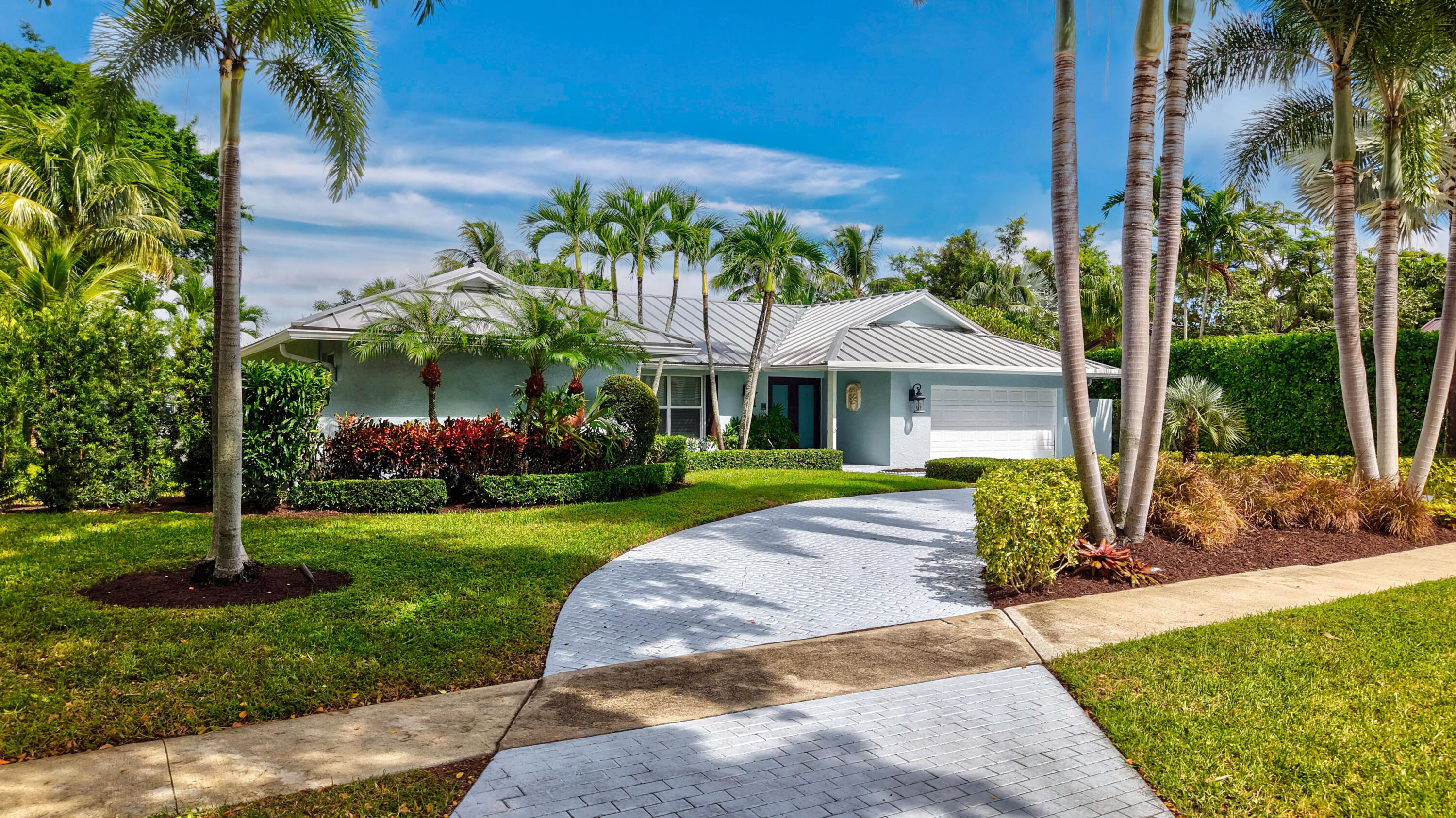 Single-story residence featuring a metal roof, paved driveway, two-car garage, and established landscaping with mature palms