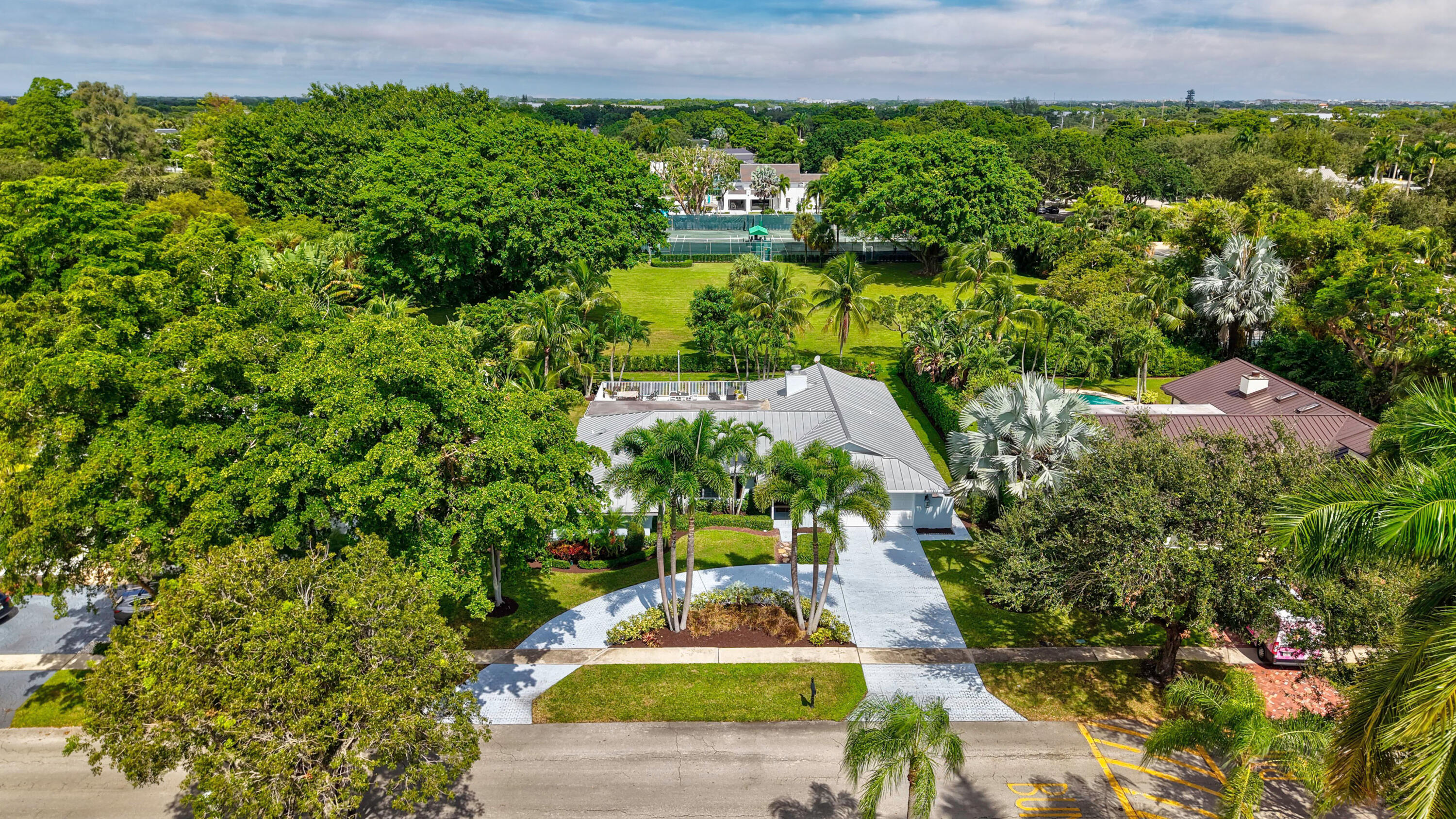 Aerial view featuring a metal roofing system, paved driveway, landscaped perimeter, and mature canopy trees