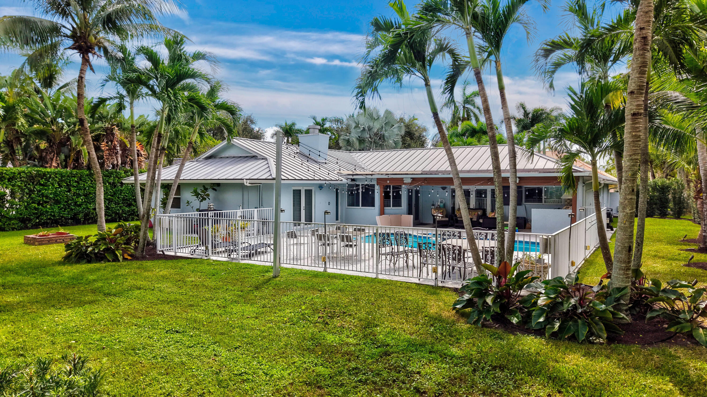 Rear exterior featuring a metal roof, a fenced-in swimming pool, a covered patio area, and mature palm trees within a landscaped yard