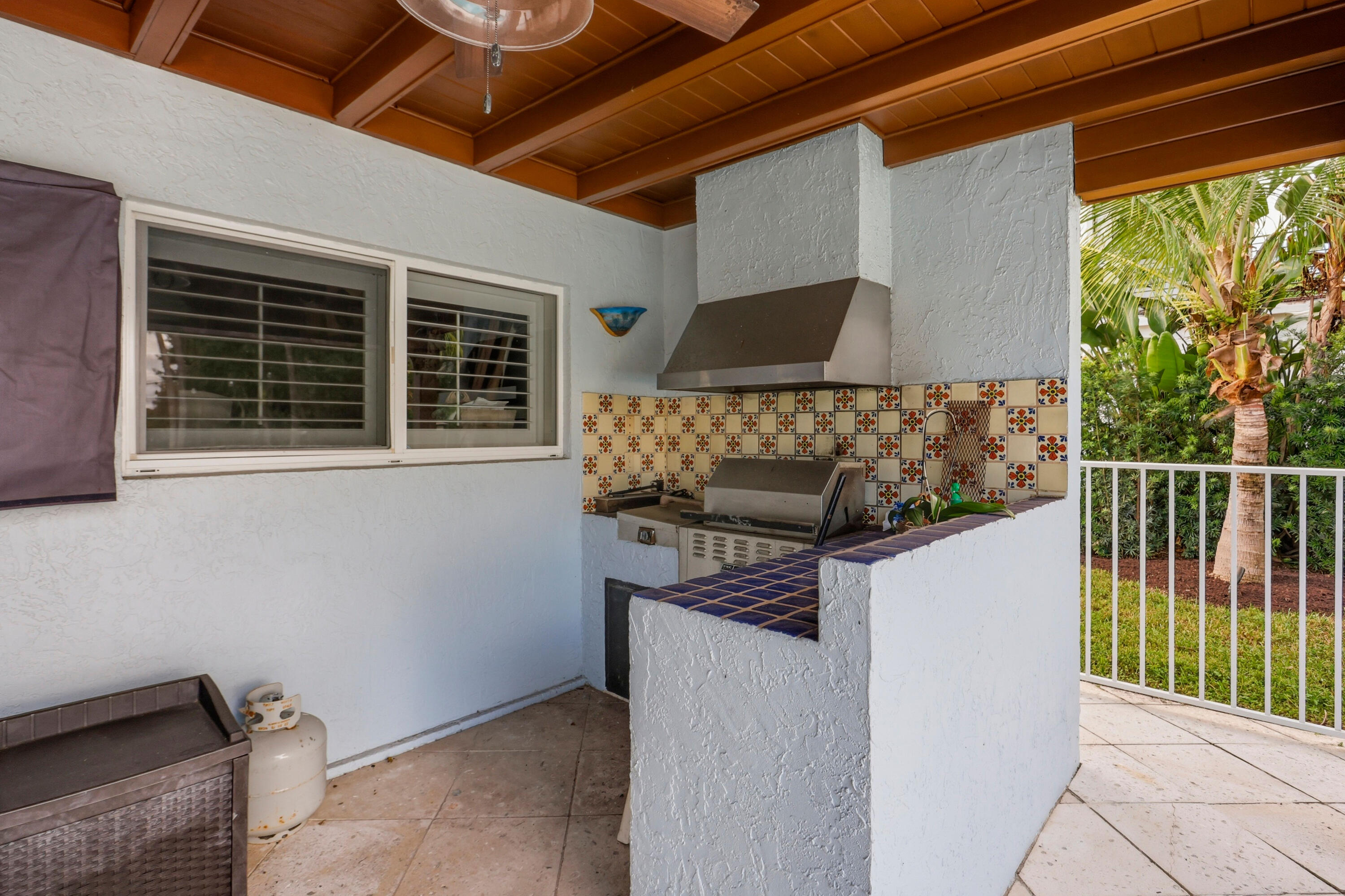 Covered outdoor kitchen featuring a built-in stainless steel grill, range hood, tiled backsplash, and tile countertop with integrated sink, situated under a wood-beam ceiling