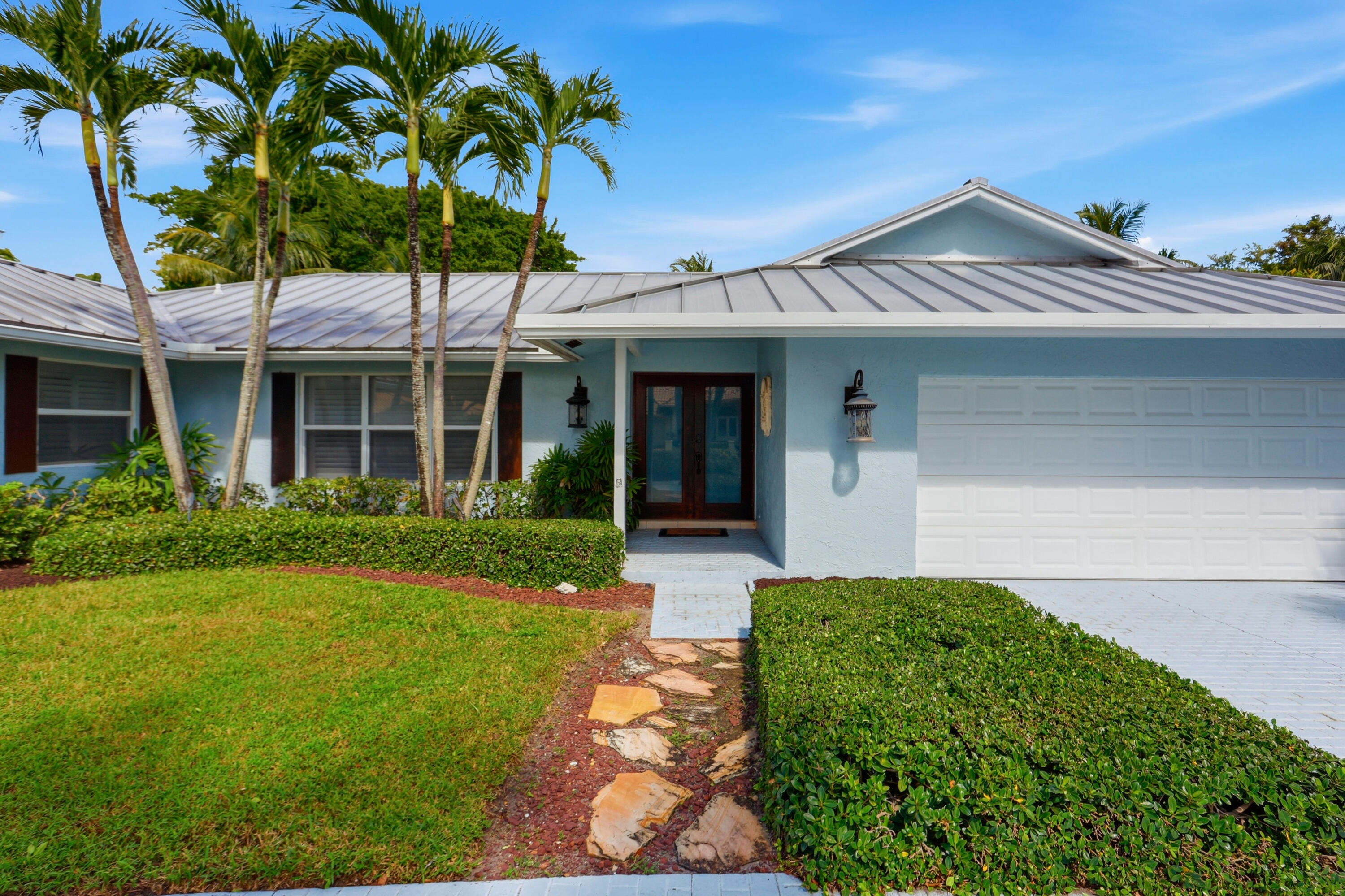 Single-story residence featuring a standing seam metal roof, stone-paved walkway, attached two-car garage, and established palm trees within the front landscape