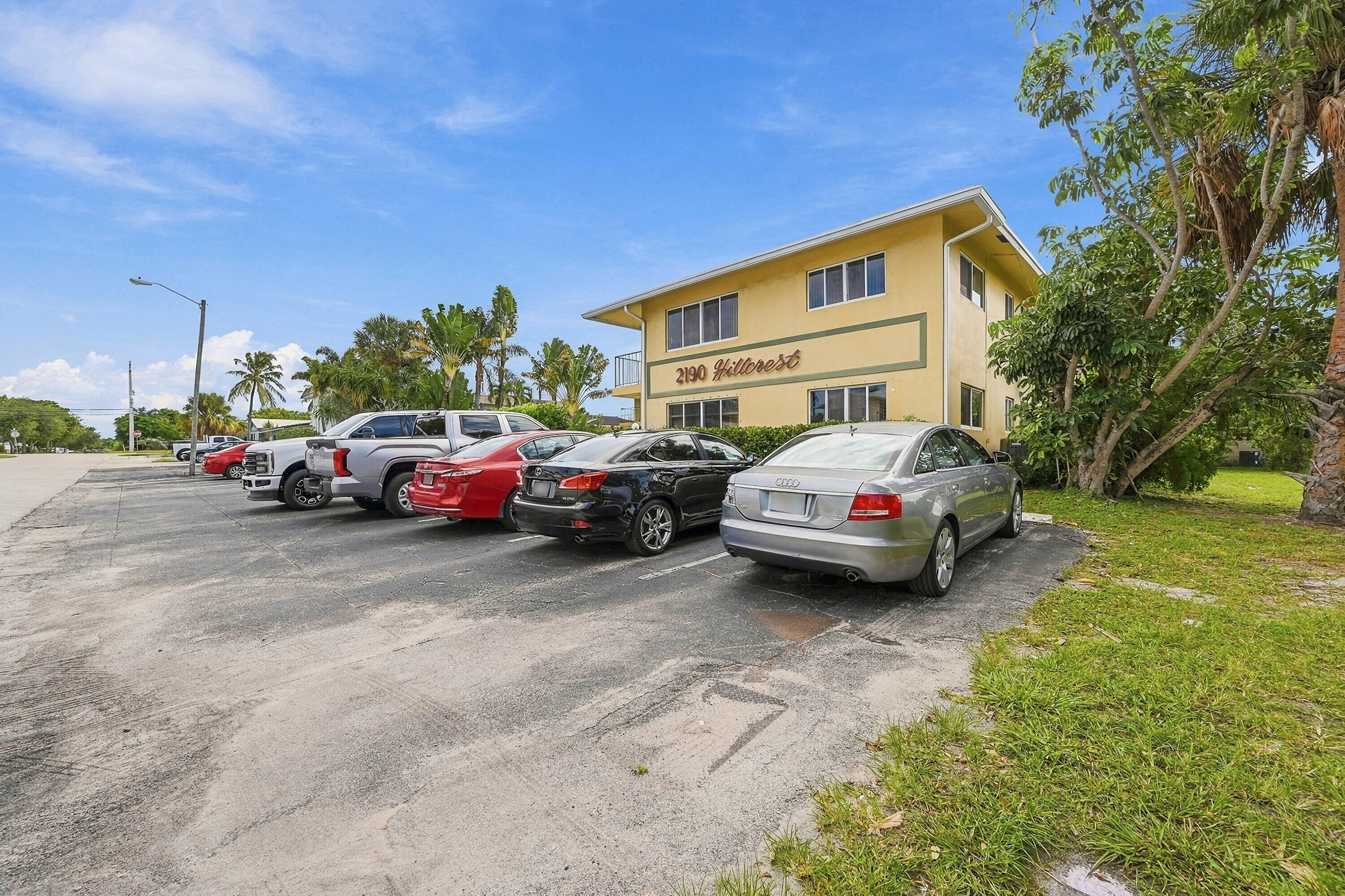 Multi-story building exterior featuring a low-pitch roofline, stucco-finish facade, designated surface parking, and mature tropical landscaping Multi-story building exterior featuring a low-pitch roofline, stucco-finish facade, designated surface parking, and mature tropical landscaping