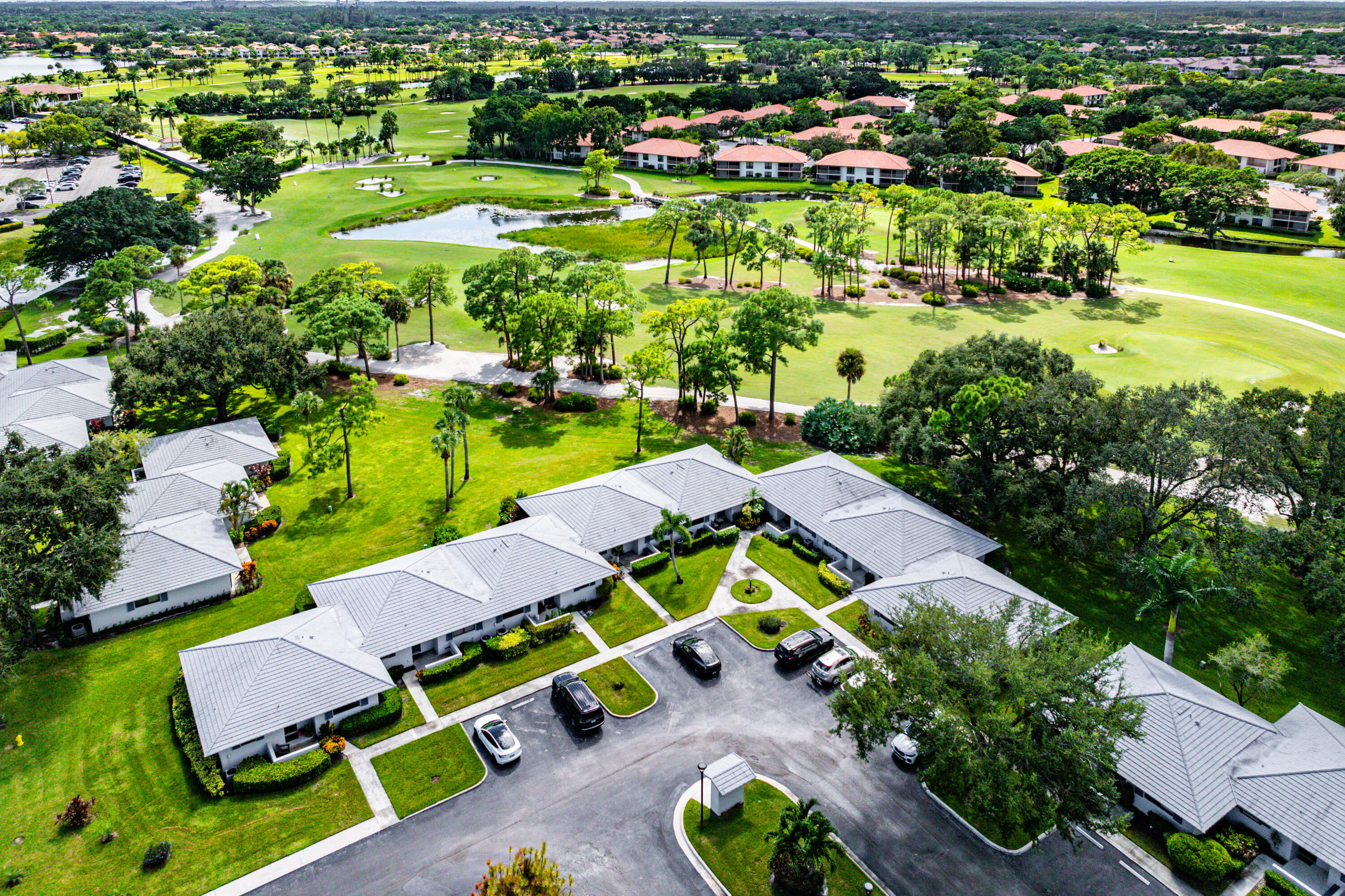 Aerial perspective of a multi-unit residential complex featuring gray tiled roofs, manicured lawns, paved parking areas, and expansive golf course grounds with water features