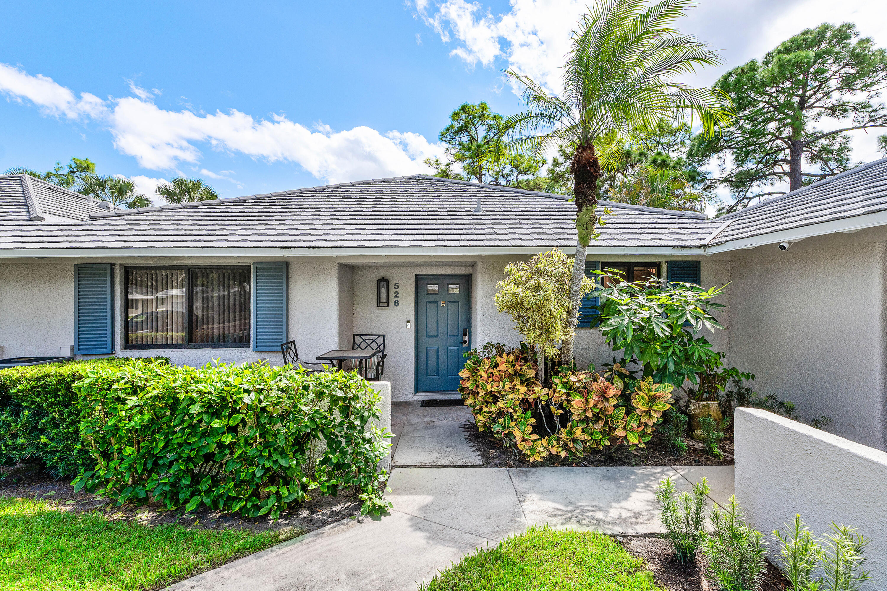Stucco exterior residence featuring a gray concrete tile roof, solid door with glass detailing, decorative blue shutters, and a paved walkway leading to the entryway
