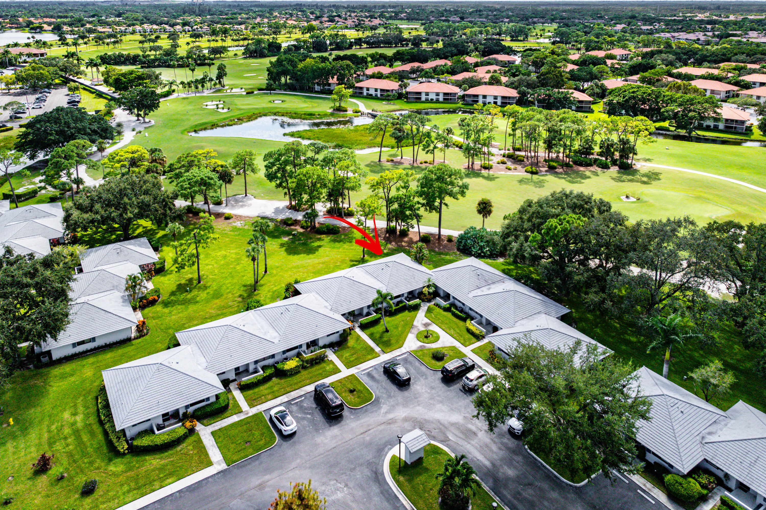 Aerial perspective of a multi-unit complex featuring light-colored shingle roofing, paved parking areas, and mature landscaping, positioned alongside a golf course with water features and expansive green space