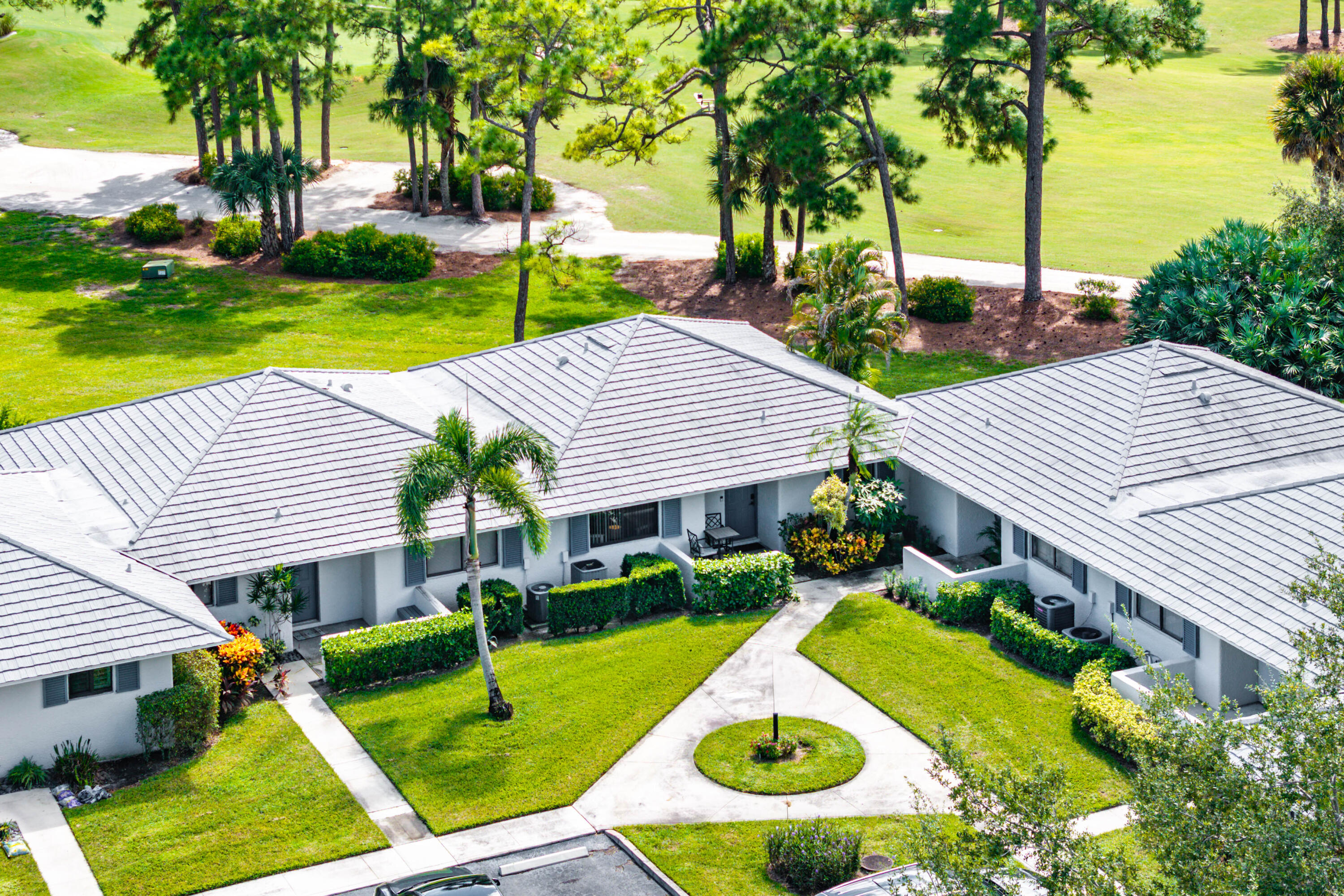 Aerial view of a residential complex featuring light-colored roof tiles, stucco siding, manicured green space, concrete walkways, and mature landscaping