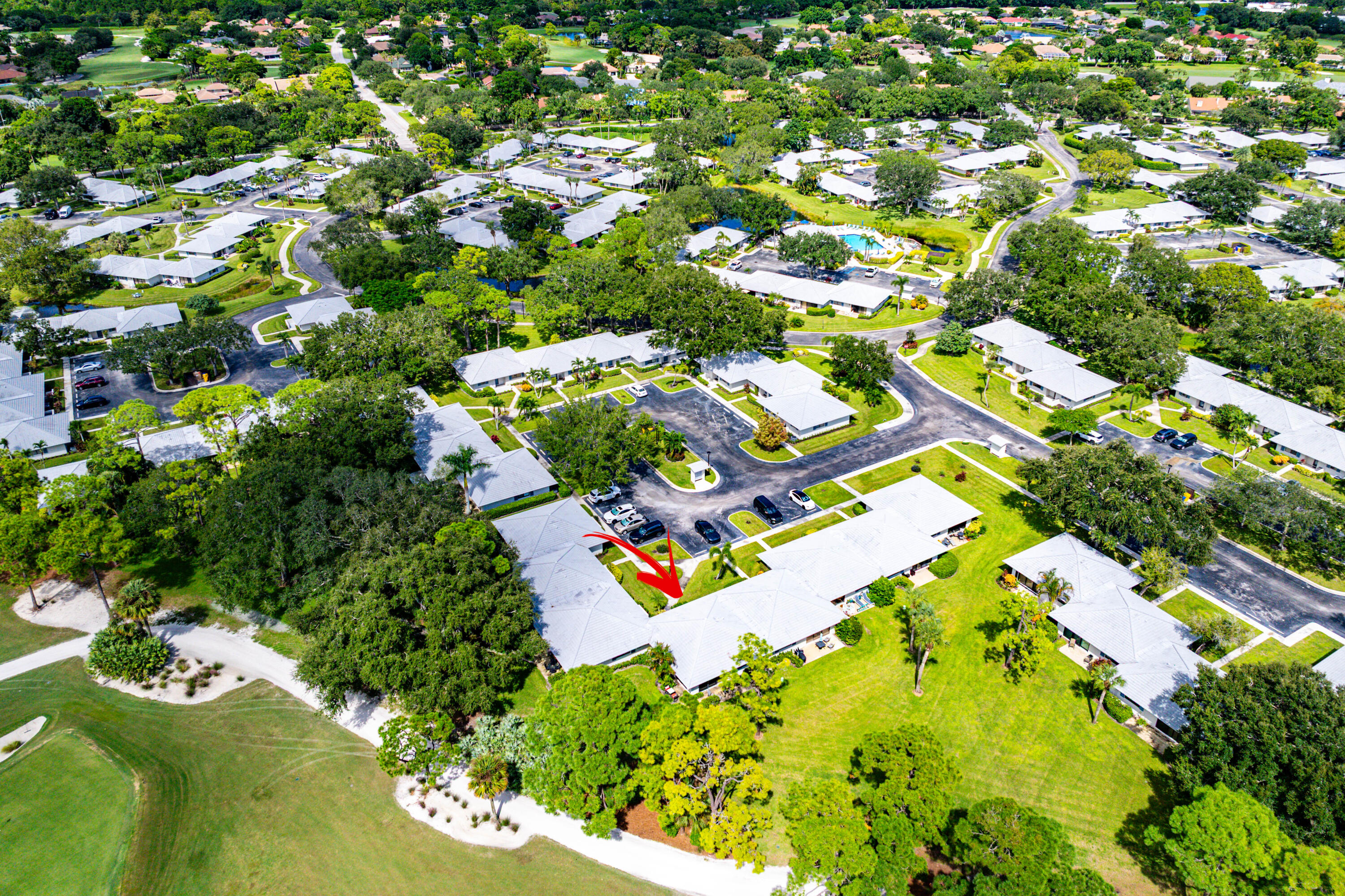 Aerial perspective of residential property featuring a light-colored roof, surrounded by mature trees, expansive green lawns, and proximity to golf course grounds