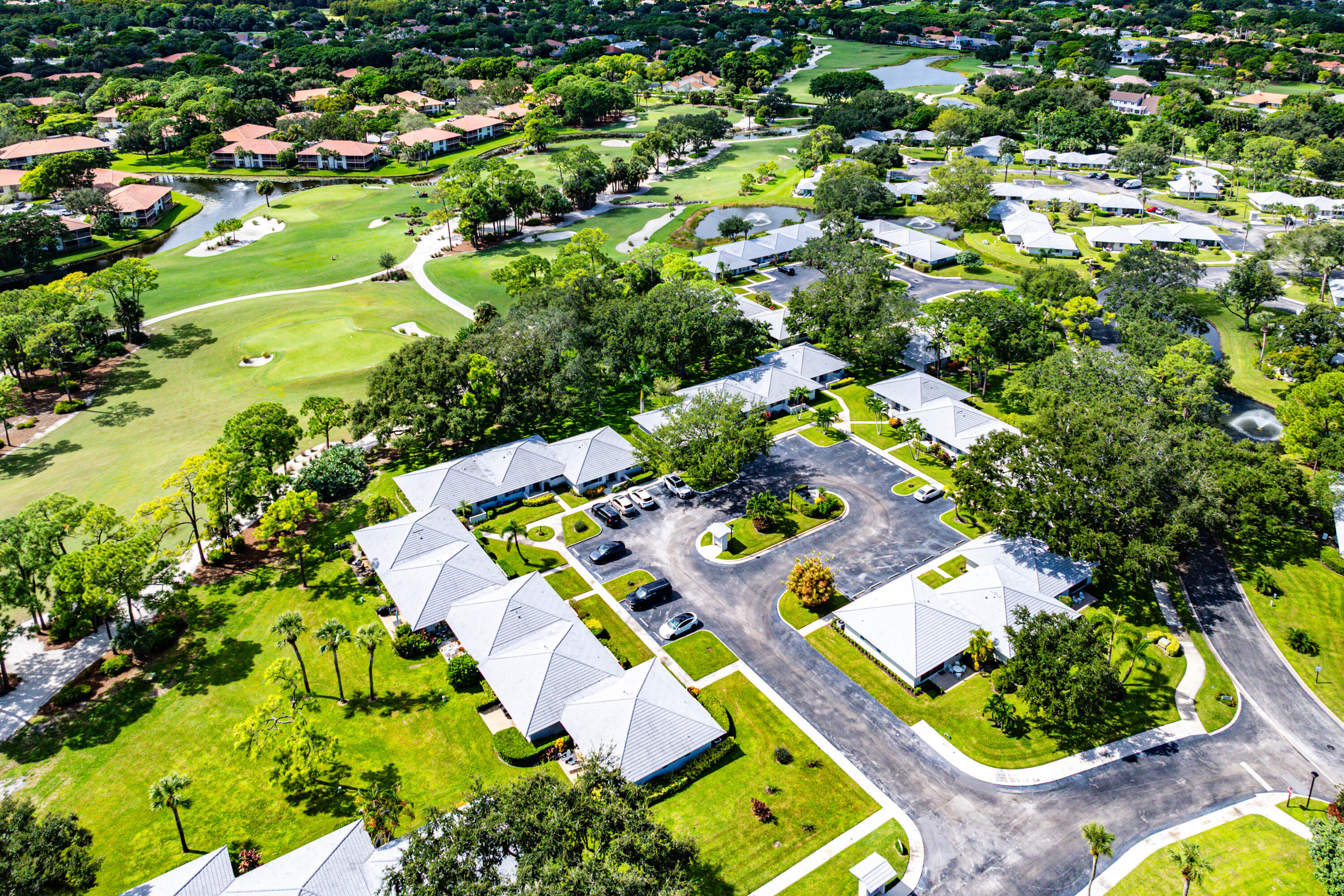 Aerial view of a residential complex featuring light-colored hip roofs, paved parking areas, extensive green space, mature trees, and direct proximity to a golf course