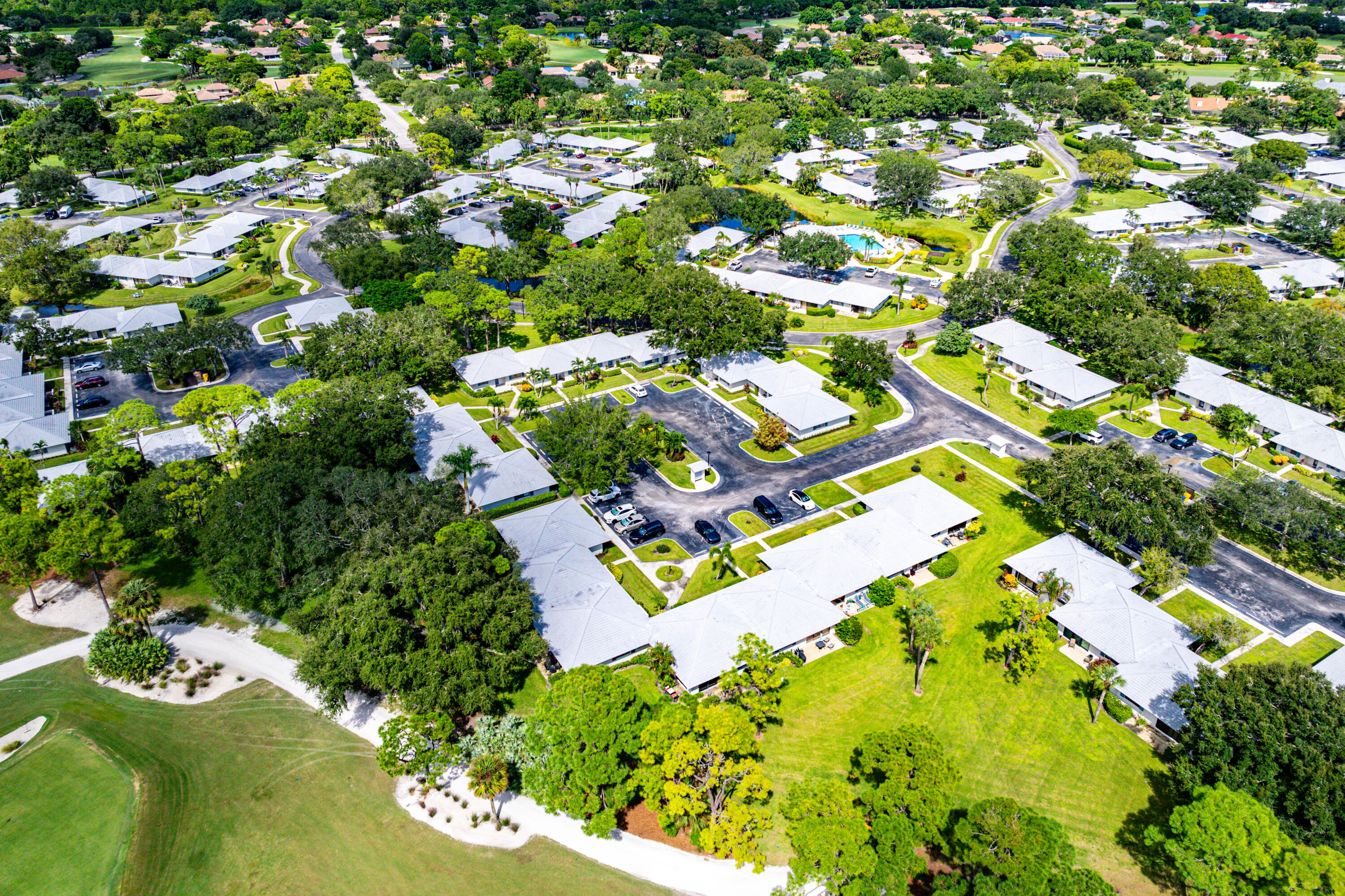 Aerial perspective featuring residential buildings with light-toned roof shingles, extensive mature tree canopy, integrated paved roadways, and landscaped green space