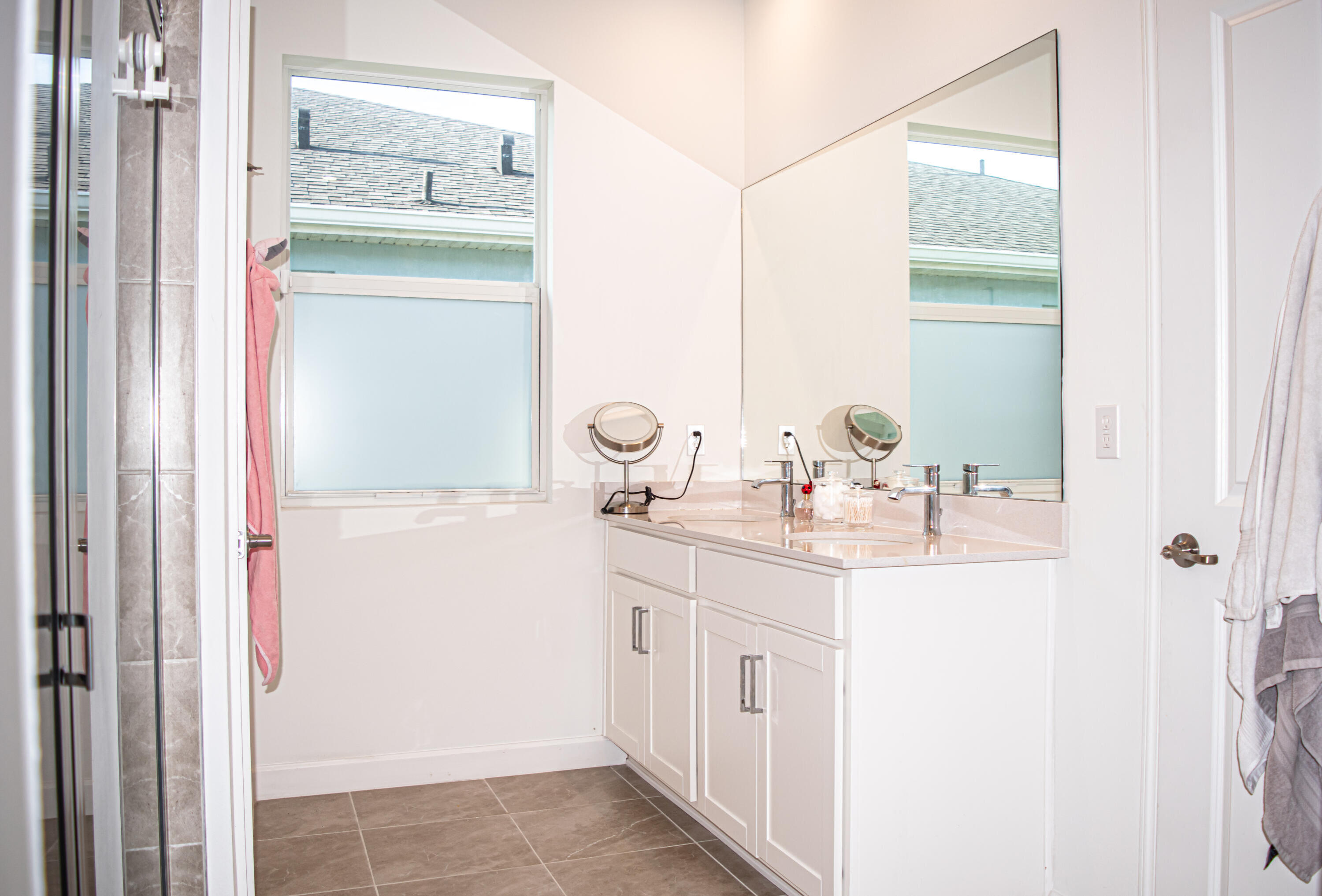 Dual-sink vanity with light-toned countertop, large wall-mounted mirror, ceramic tile flooring, and frosted window