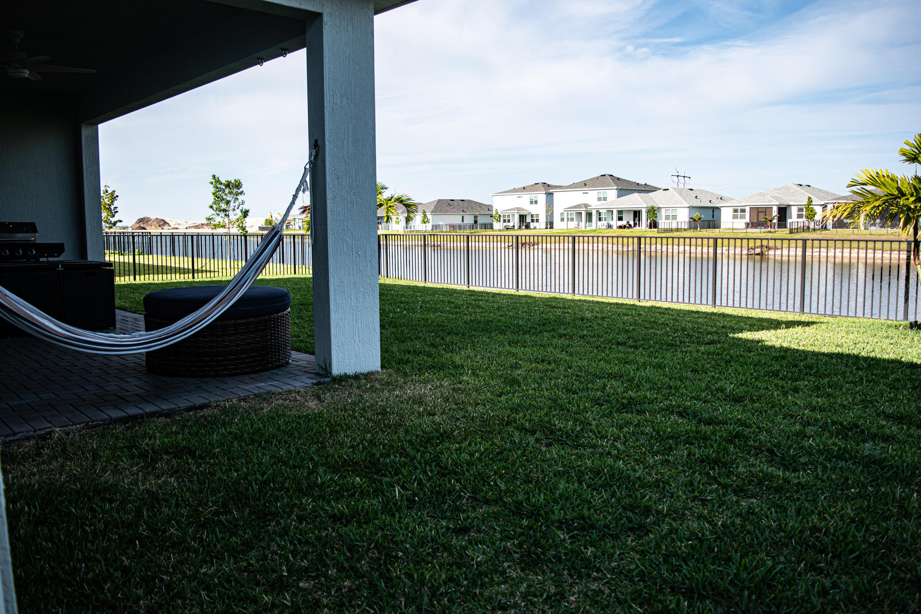 Covered patio with paver flooring, manicured lawn, and aluminum perimeter fencing bordering a waterfront