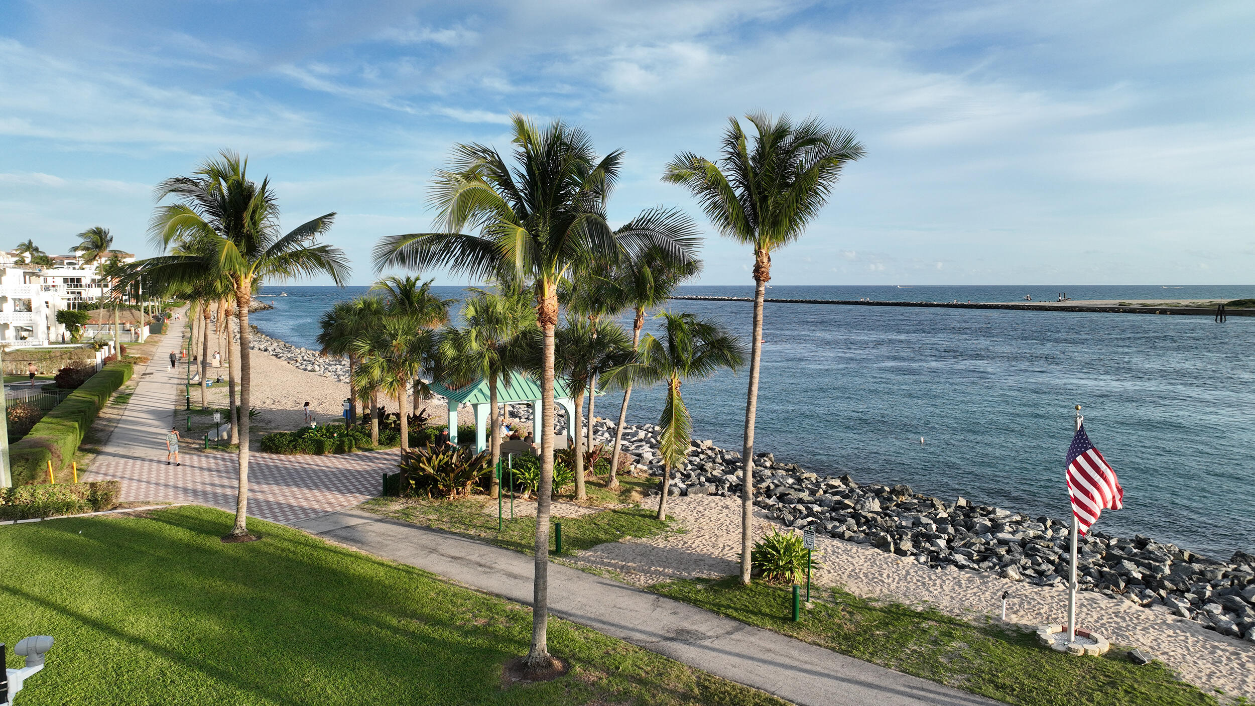 Waterfront property featuring a paved promenade, mature palm trees, expansive lawn, and a rock-lined shoreline
