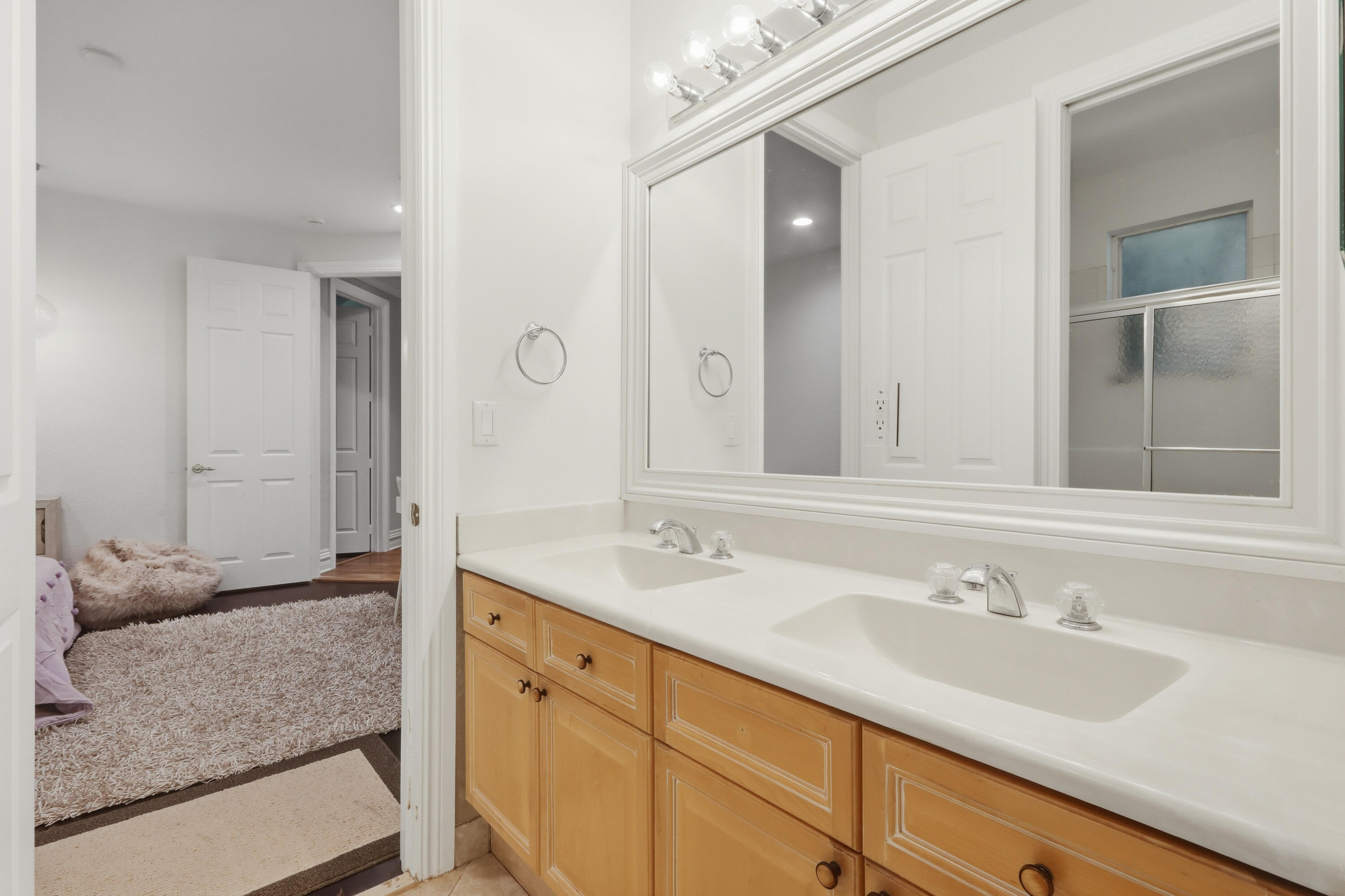 Double vanity featuring natural wood cabinetry, integrated white countertops with dual sinks, chrome fixtures, and a large framed wall mirror with integrated lighting