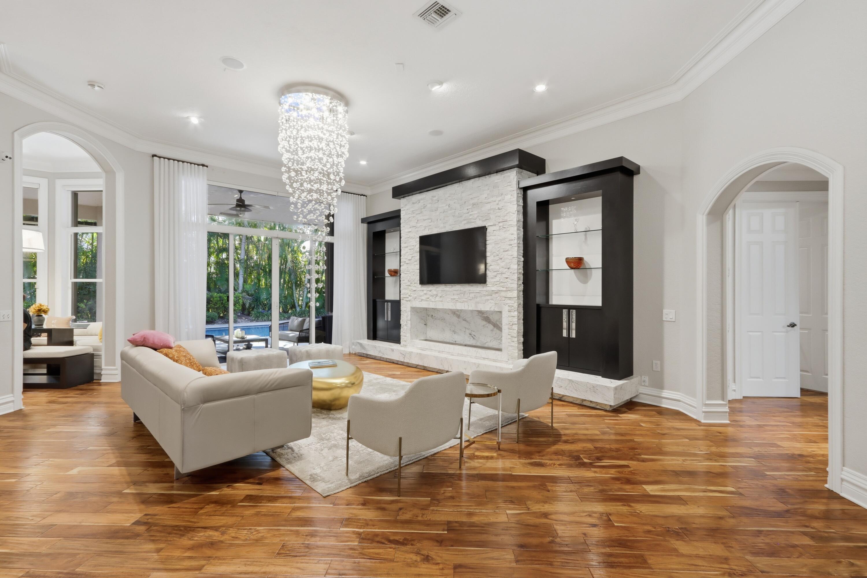 Formal living space featuring wood-finish flooring, a floor-to-ceiling stone-textured fireplace with built-in dark wood cabinetry, an intricate crystal chandelier, and arched entryways Formal living space featuring wood-finish flooring, a floor-to-ceiling stone-textured fireplace with built-in dark wood cabinetry, an intricate crystal chandelier, and arched entryways