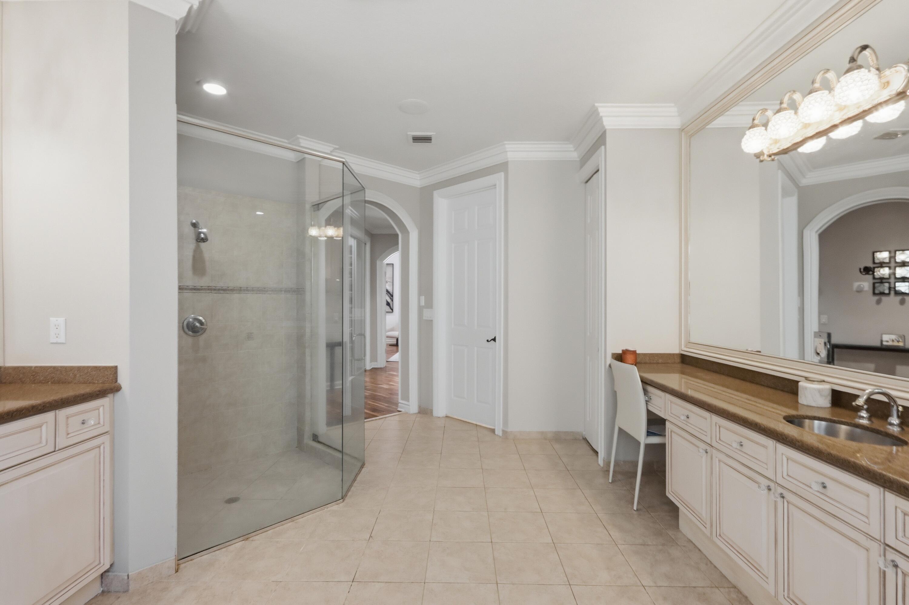 Glass-enclosed corner shower with neutral tile surround, extensive built-in cabinetry with stone-finish countertops, ceramic tile flooring, crown molding, and an oversized wall-mounted mirror