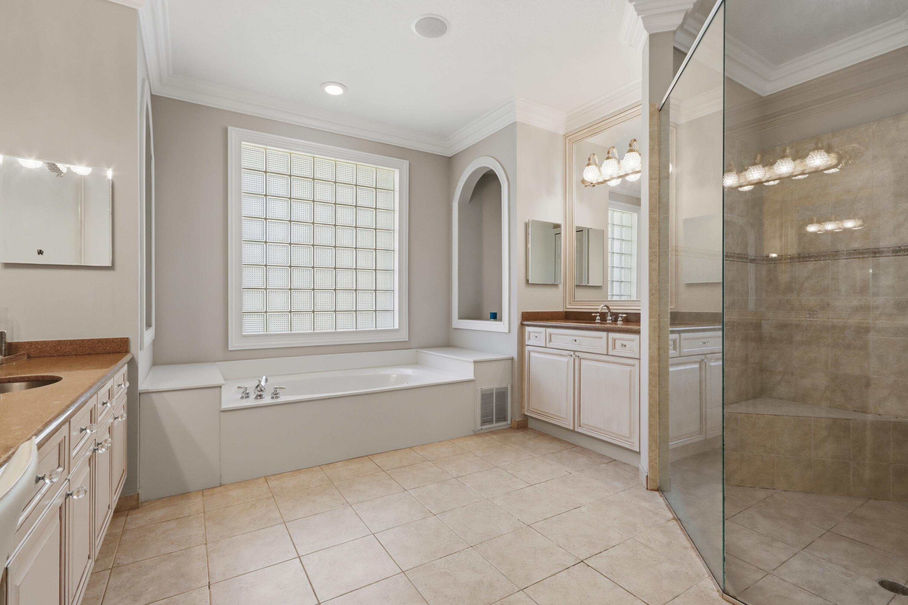 Bathroom featuring a soaking tub, glass block window, stone-tiled walk-in shower with bench, dual vanities with stone countertops, and arched wall niche