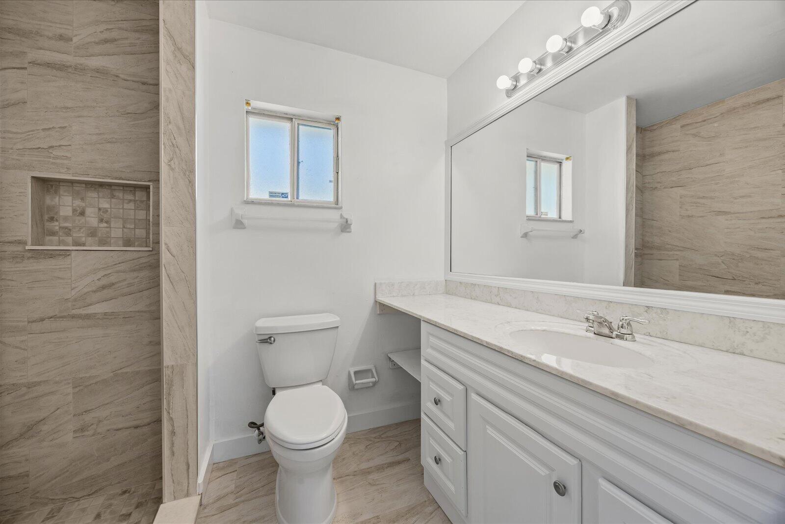 Bathroom featuring a vanity with stone-finish countertop, full-width wall mirror, wood-finish flooring, and a shower enclosure with tile surround and built-in recessed shelving