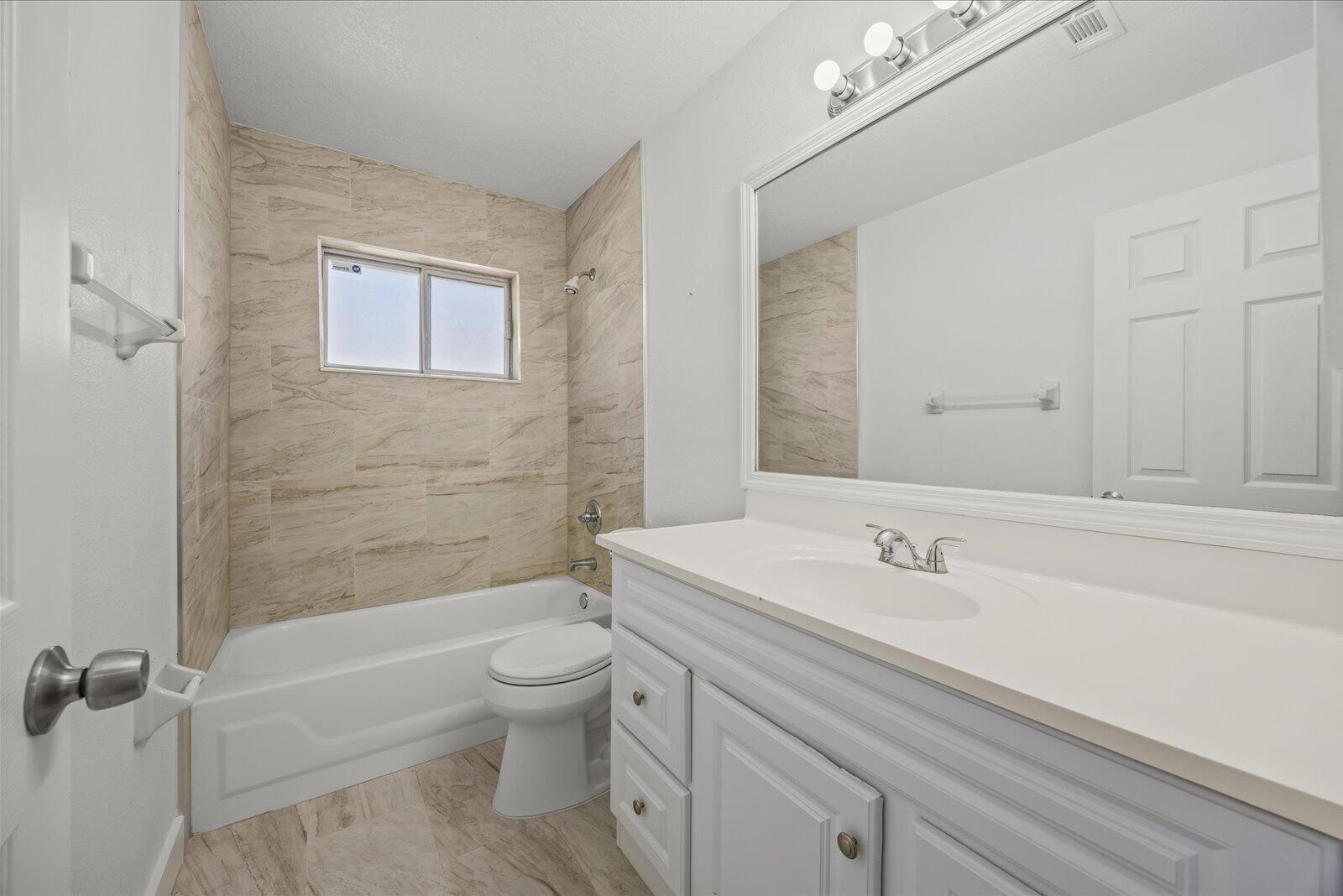 Bathroom featuring a white vanity with an integrated basin, large wall-mounted mirror, three-bulb vanity lighting, and a tub-shower combination enclosed by neutral-toned tiled walls