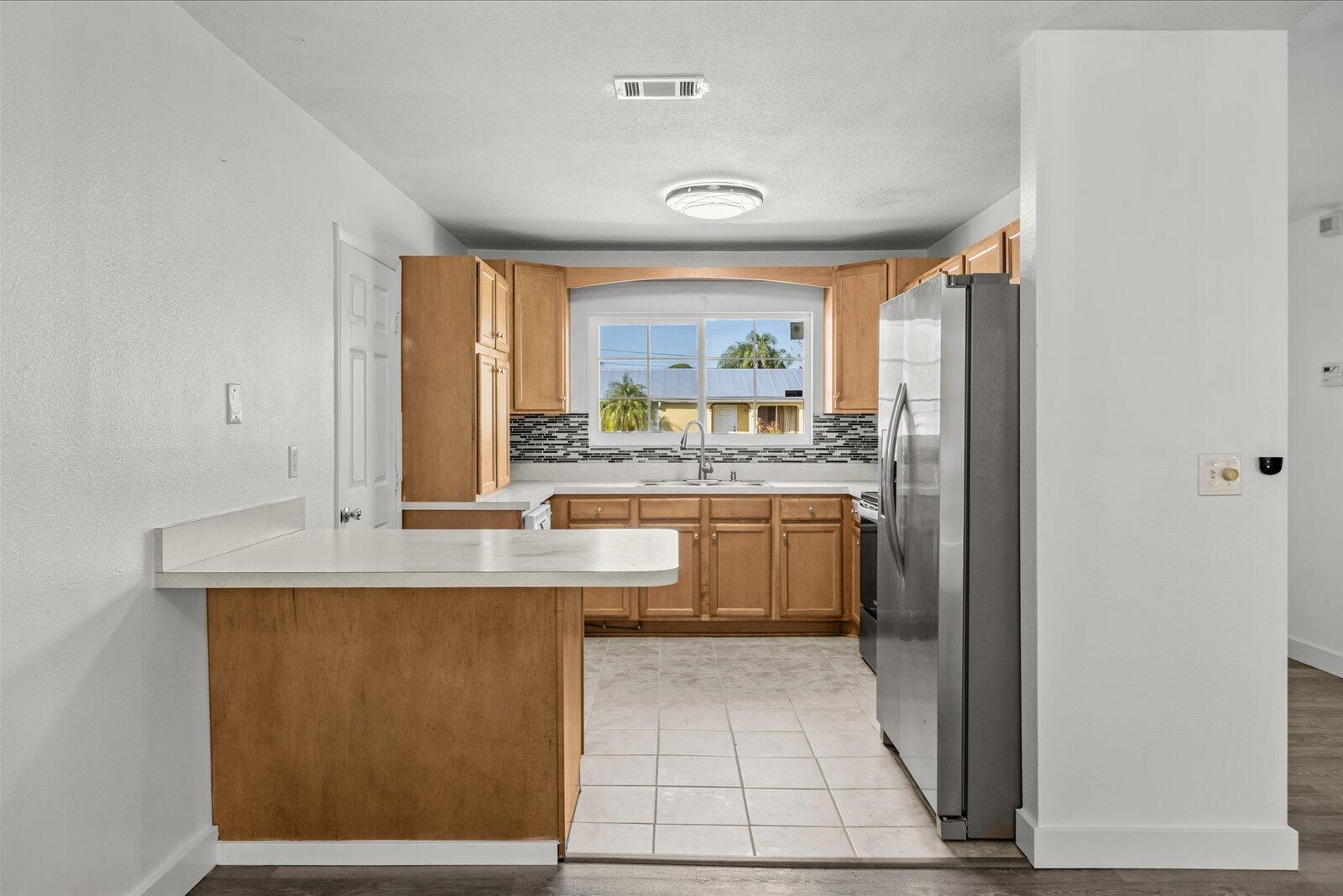 Kitchen featuring wood cabinetry, stone-style tile backsplash, white countertops, tile flooring, and stainless steel refrigerator
