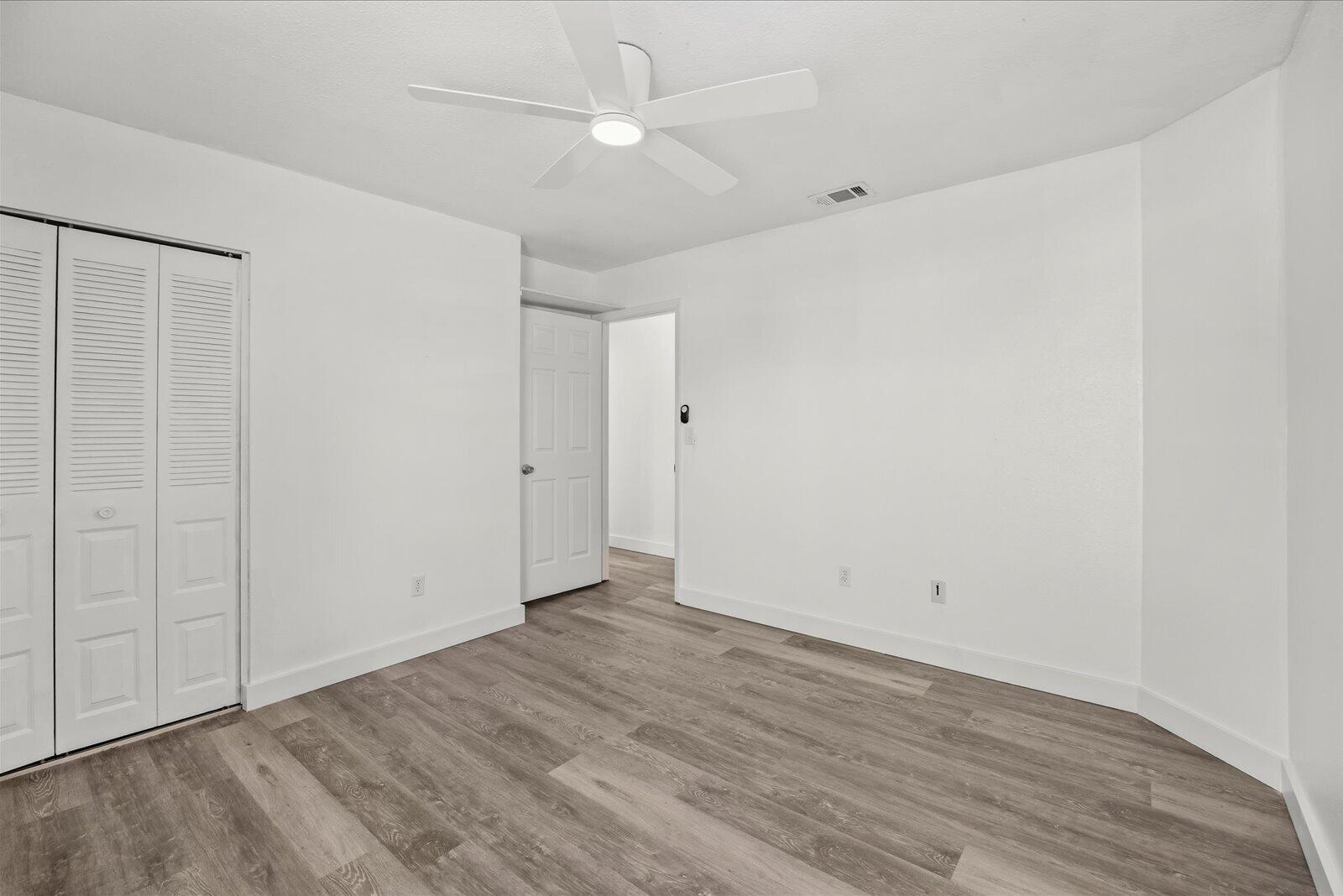 Neutral-toned room featuring wood-finish flooring, a white five-blade ceiling fan with integrated lighting, and louvered double-door closet cabinetry