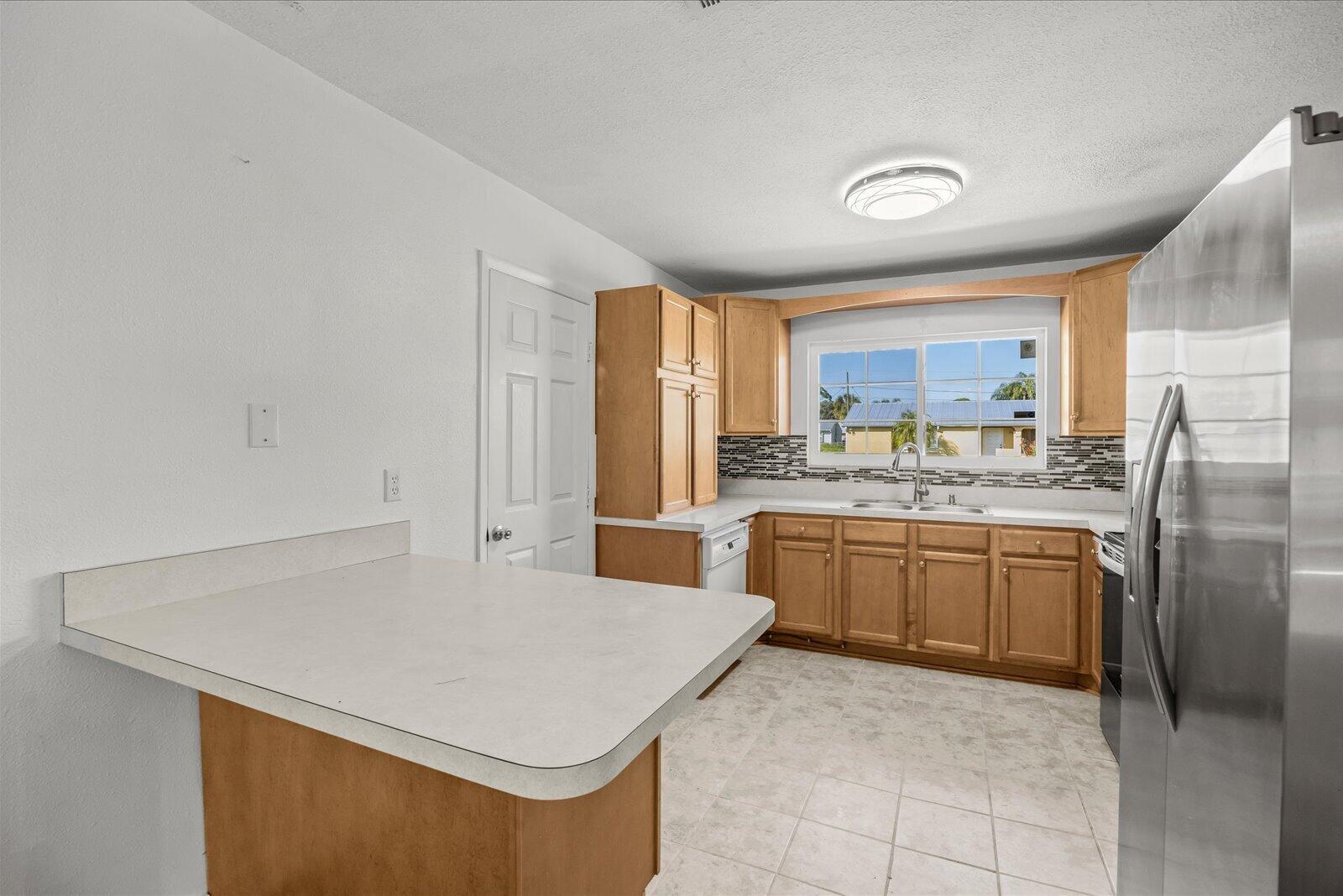 Kitchen featuring wood cabinetry, solid-surface countertops, tile backsplash, stainless steel refrigerator, and tiled flooring