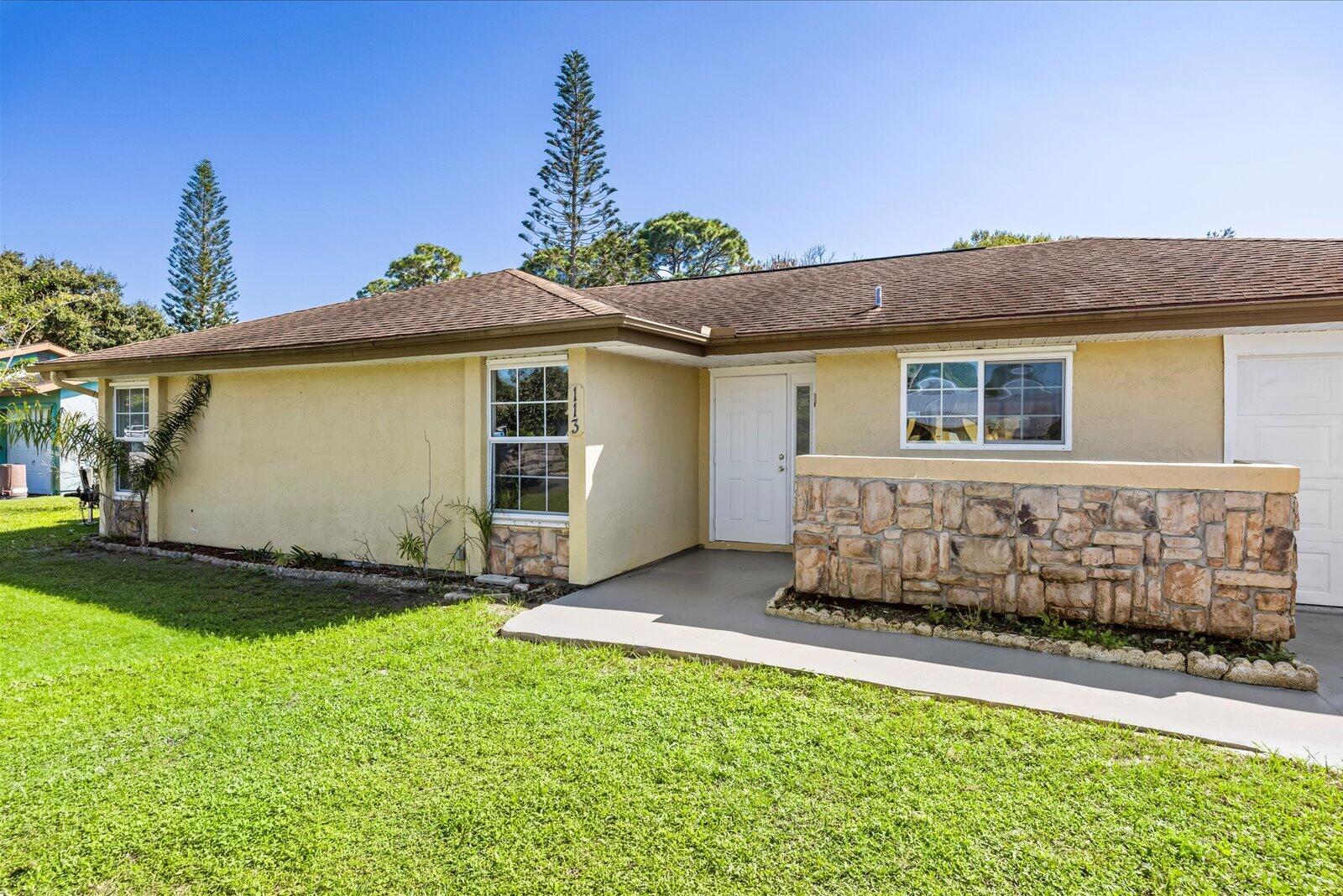 Single-story exterior featuring light-toned stucco, a multi-paneled front door, stone masonry facade detail, and a concrete walkway